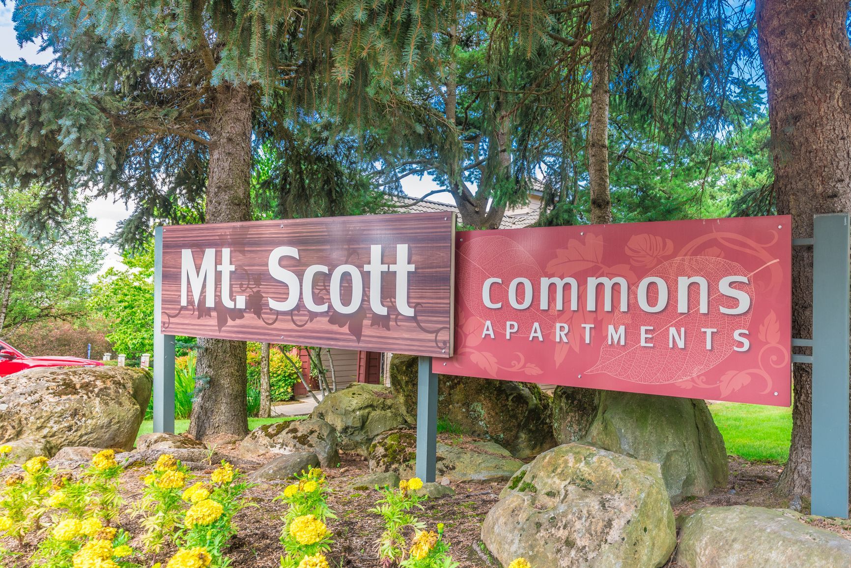 A sign for mt. scott commons apartments is surrounded by trees and flowers.