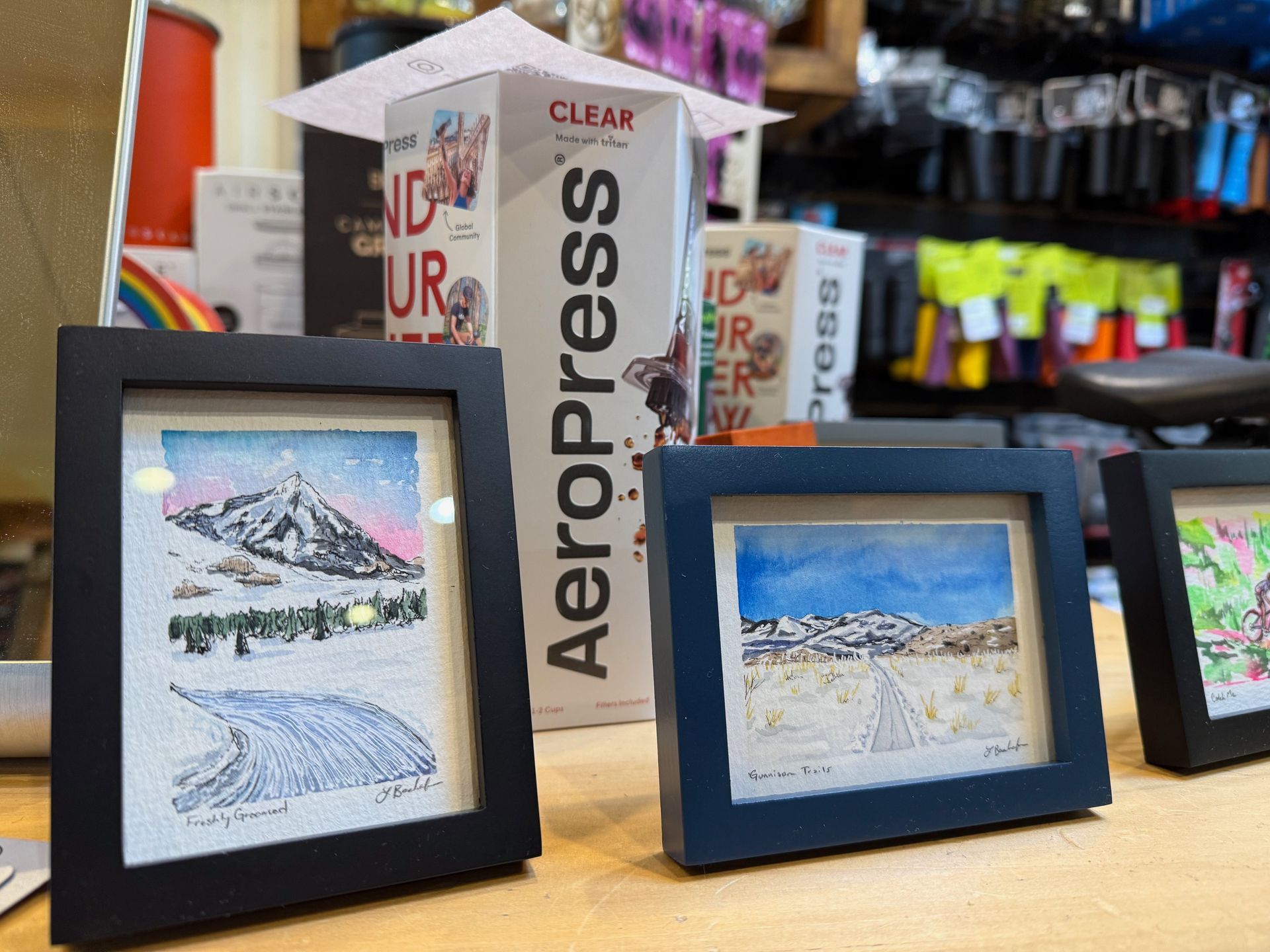 Three framed pictures are on a table in front of a box that says aeropress