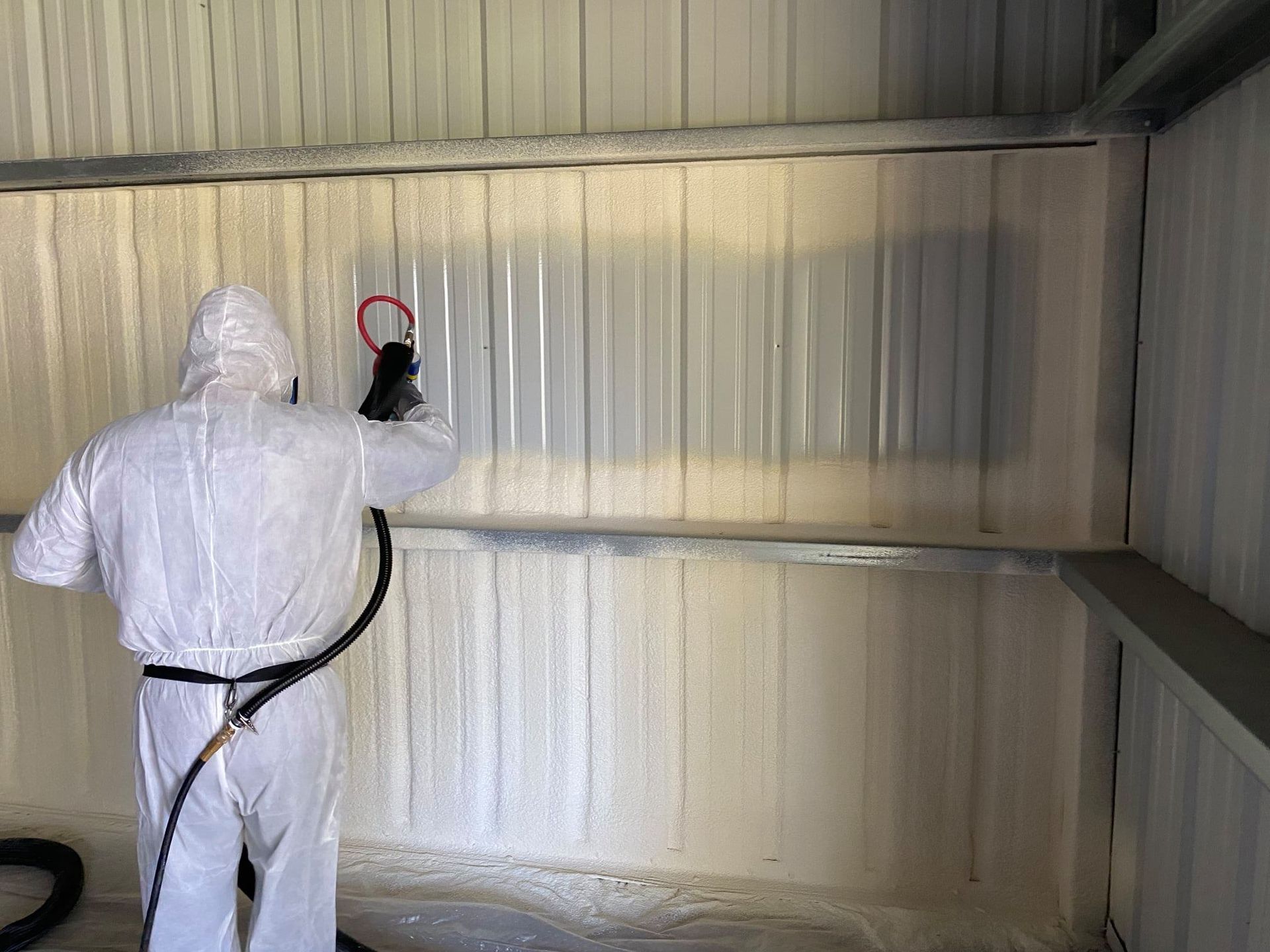 Person in protective suit spraying foam insulation on a metal wall inside a building.