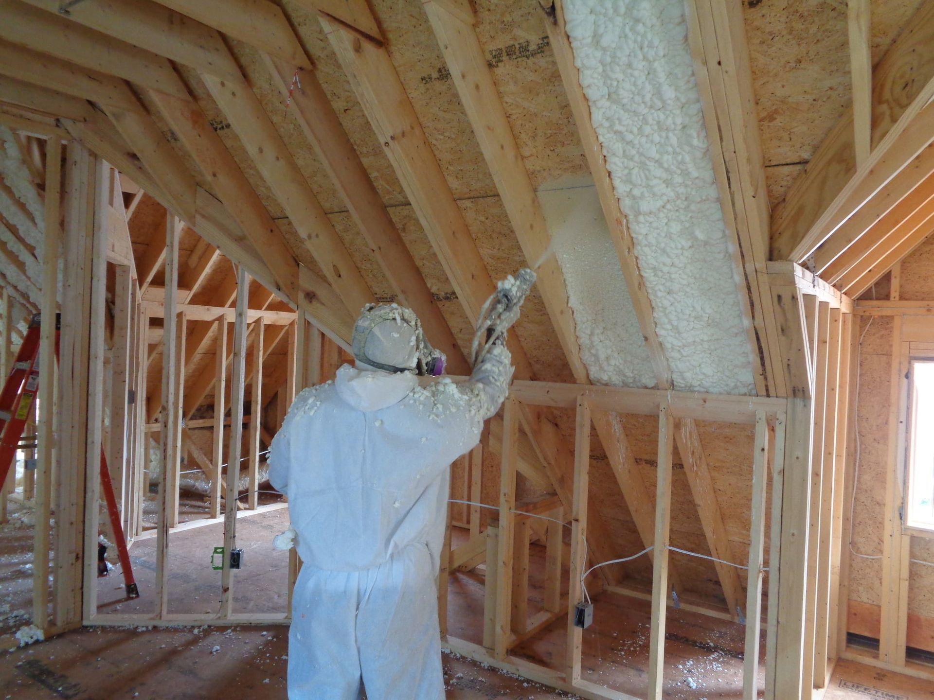 Worker in protective suit spraying insulation foam onto rafters in a wooden-framed building.