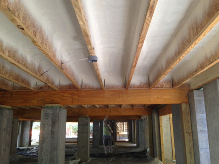 Underside of a house under construction; wooden beams and concrete pillars support a spray-foam insulated ceiling. A person stands below.
