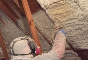 Person spraying foam insulation in an attic, wearing protective gear.