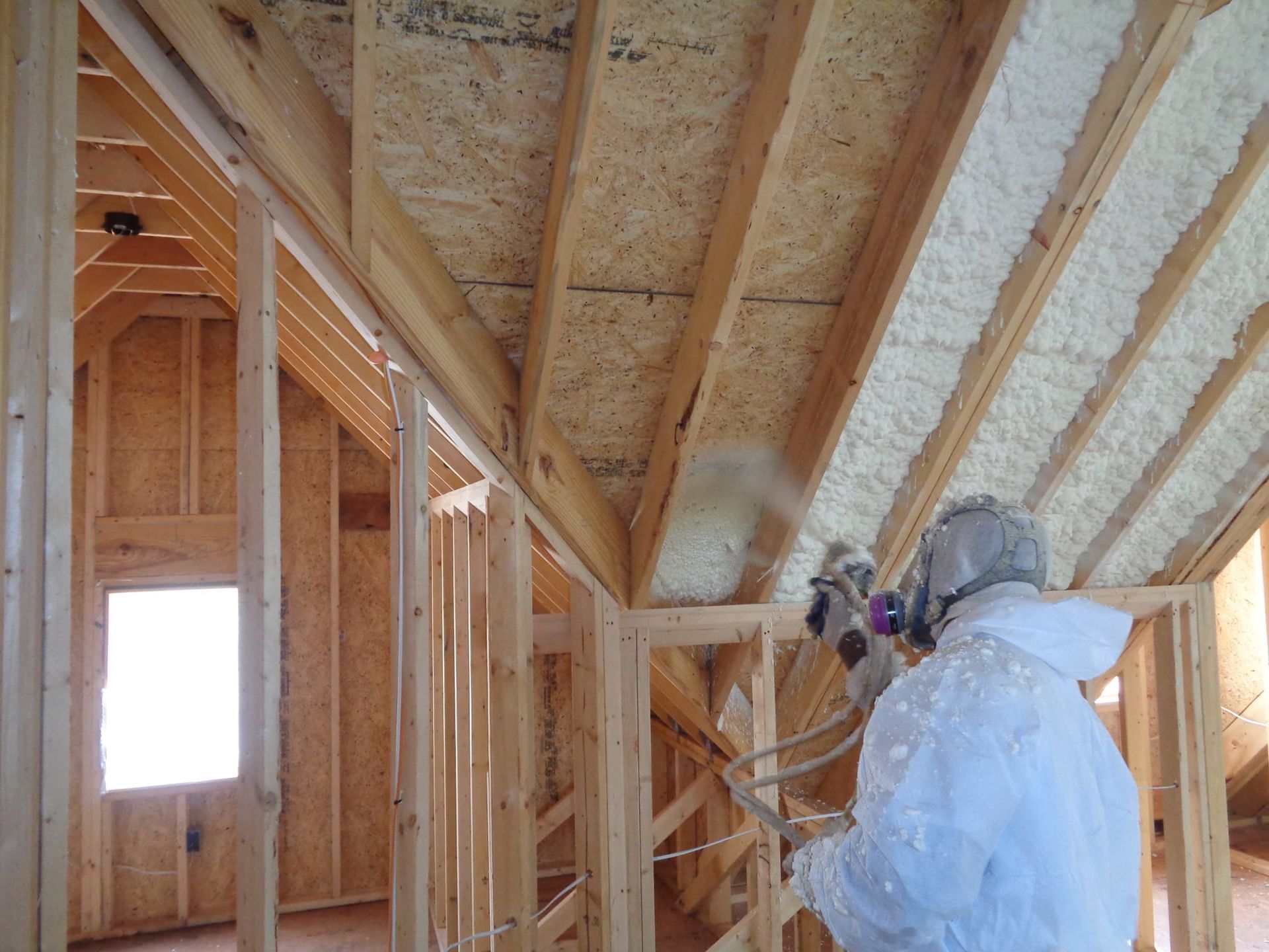 Person spraying foam insulation in a wooden structure; protective gear visible.