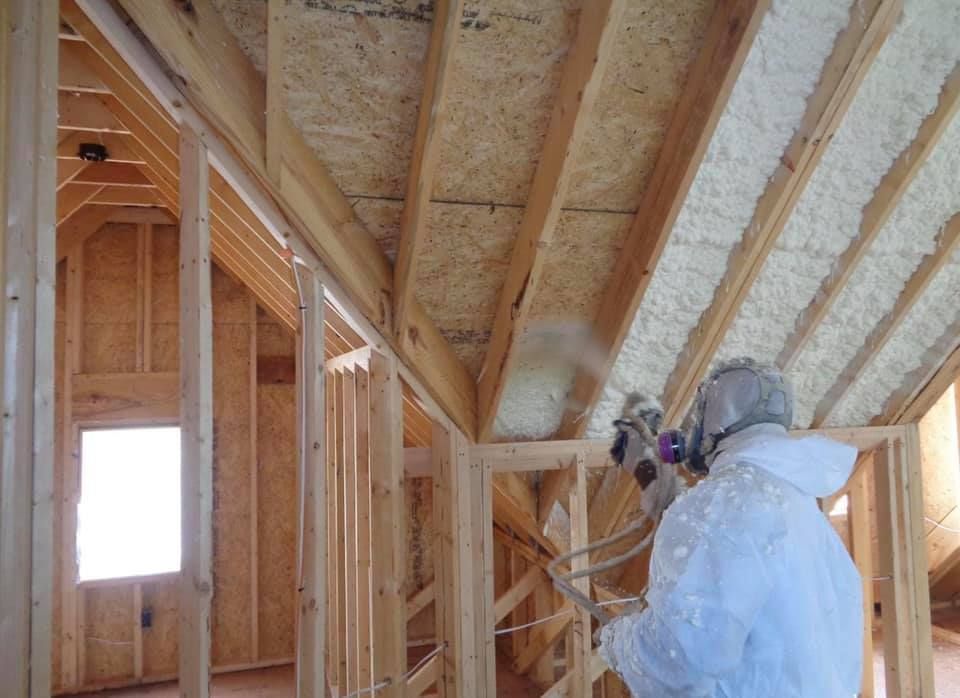 Person in protective suit spraying foam insulation into wooden attic frame.