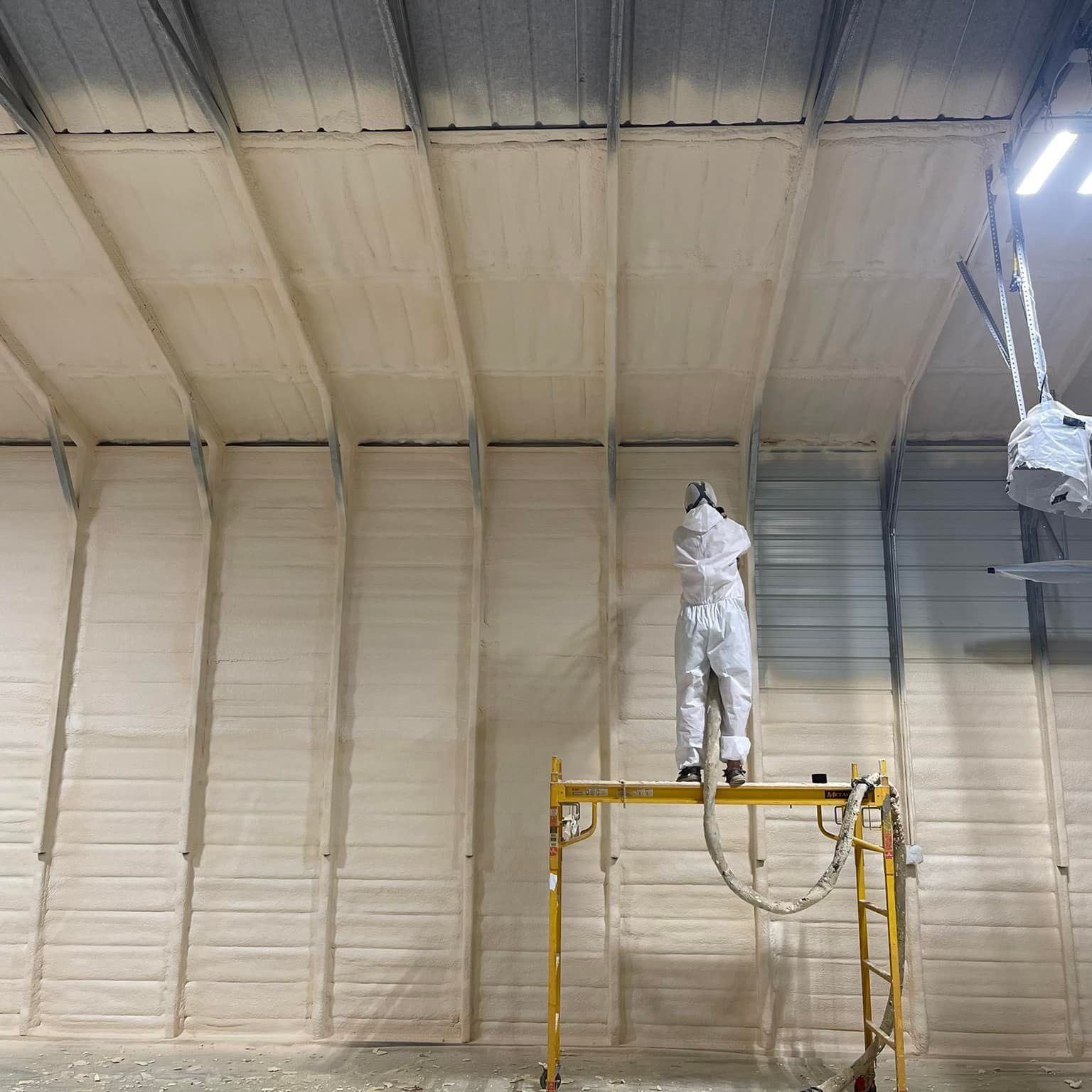 Person in protective suit spraying foam insulation on a warehouse wall and ceiling.