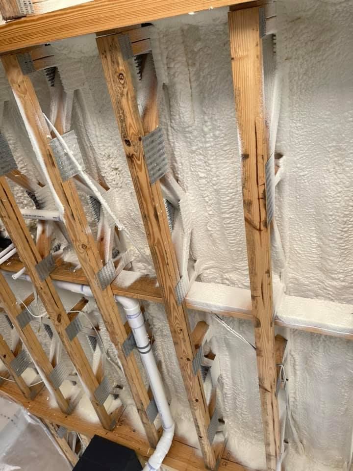 Wooden joists and plumbing pipes against a wall insulated with white spray foam.