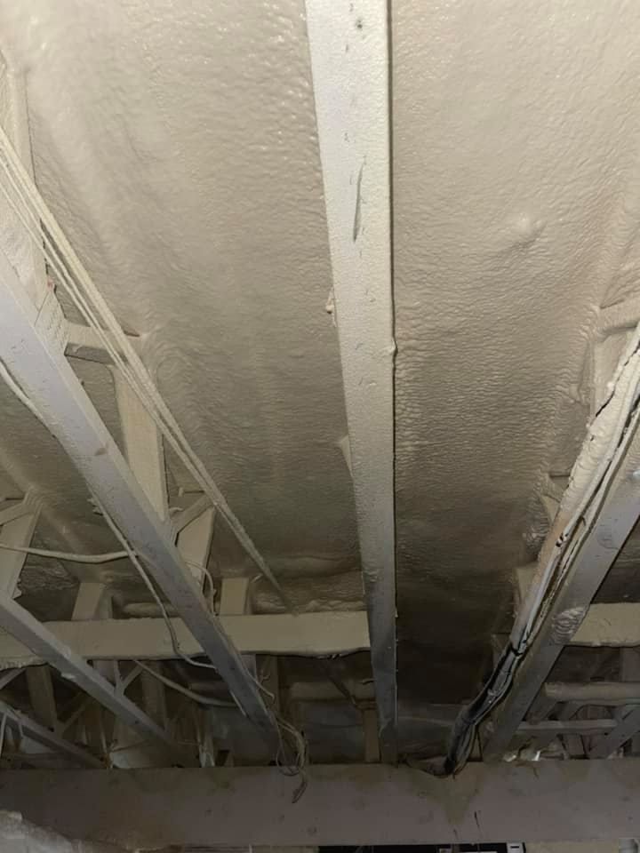 Spray foam insulation applied to the underside of a ceiling with exposed beams. White and light beige hues.