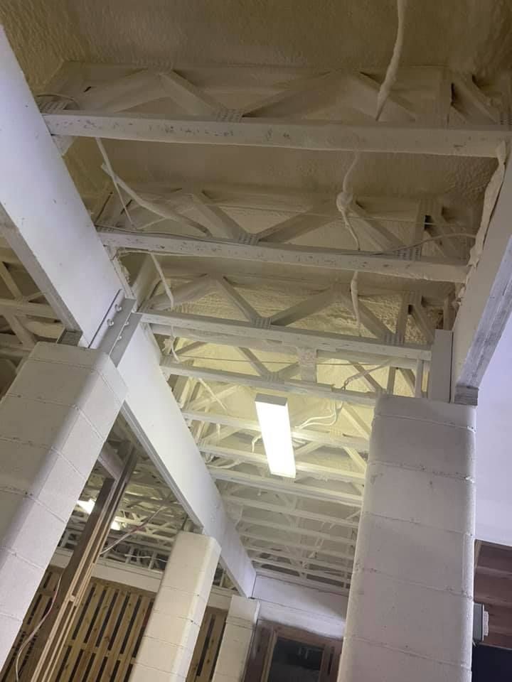White-painted beams and trusses in a ceiling, with white foam insulation visible. A fluorescent light hangs.
