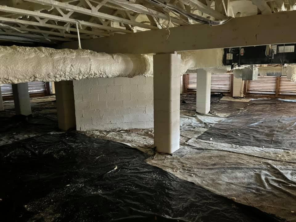 Crawlspace with spray foam insulation on beams and pipes, black plastic ground cover.