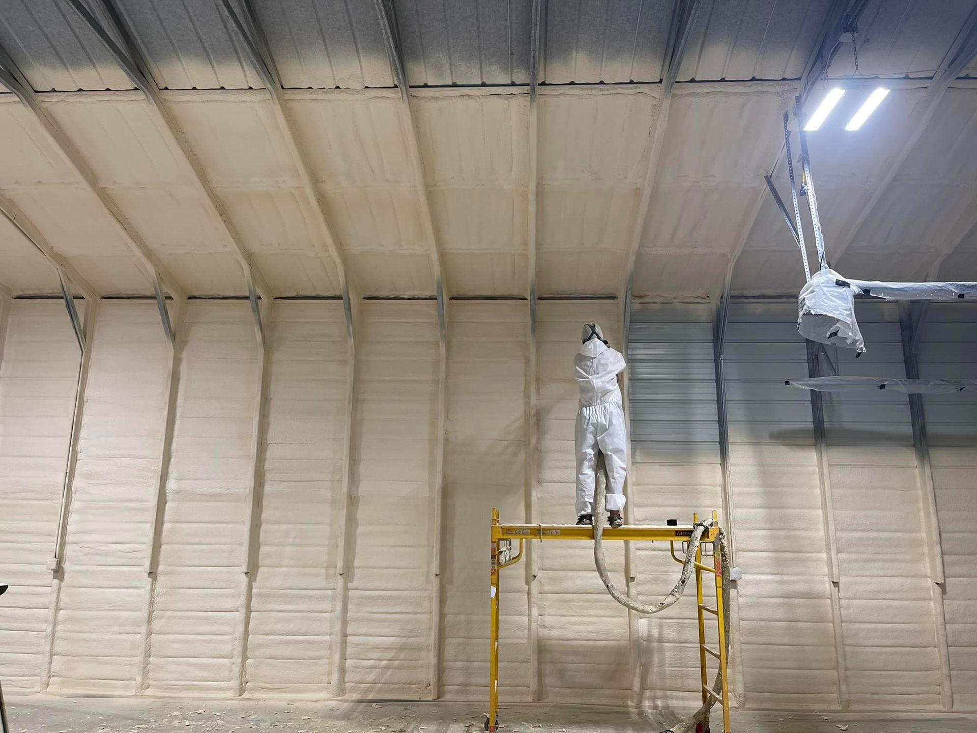 Person in protective suit spraying insulation on a warehouse wall and ceiling.