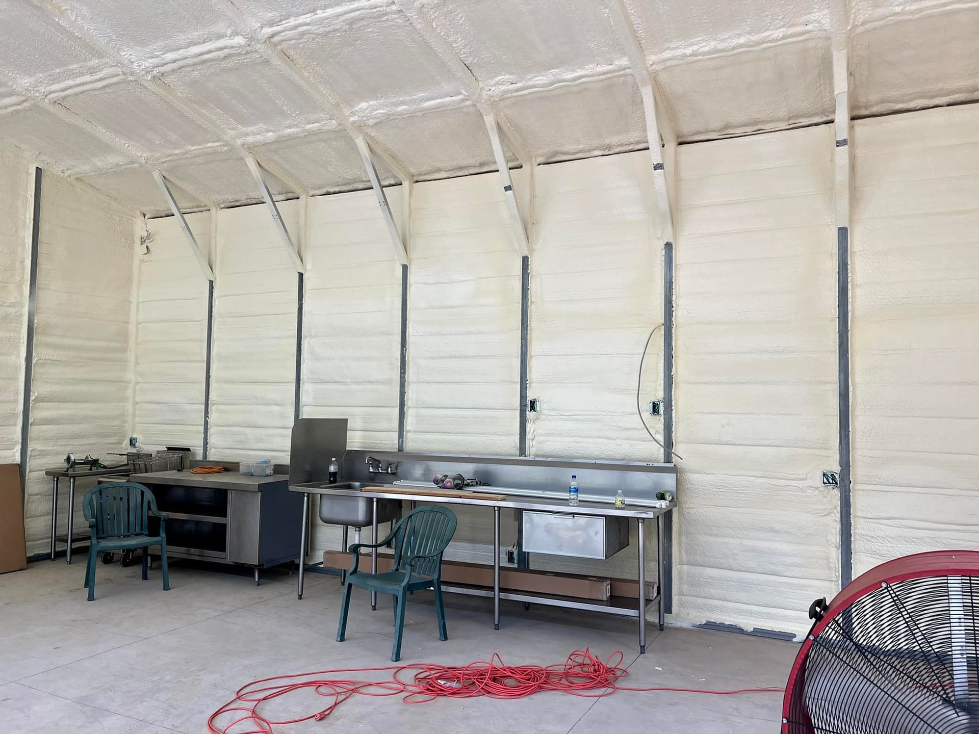 Interior of a building with foam insulation on walls and ceiling. Contains work surfaces and chairs; red cord on the floor.