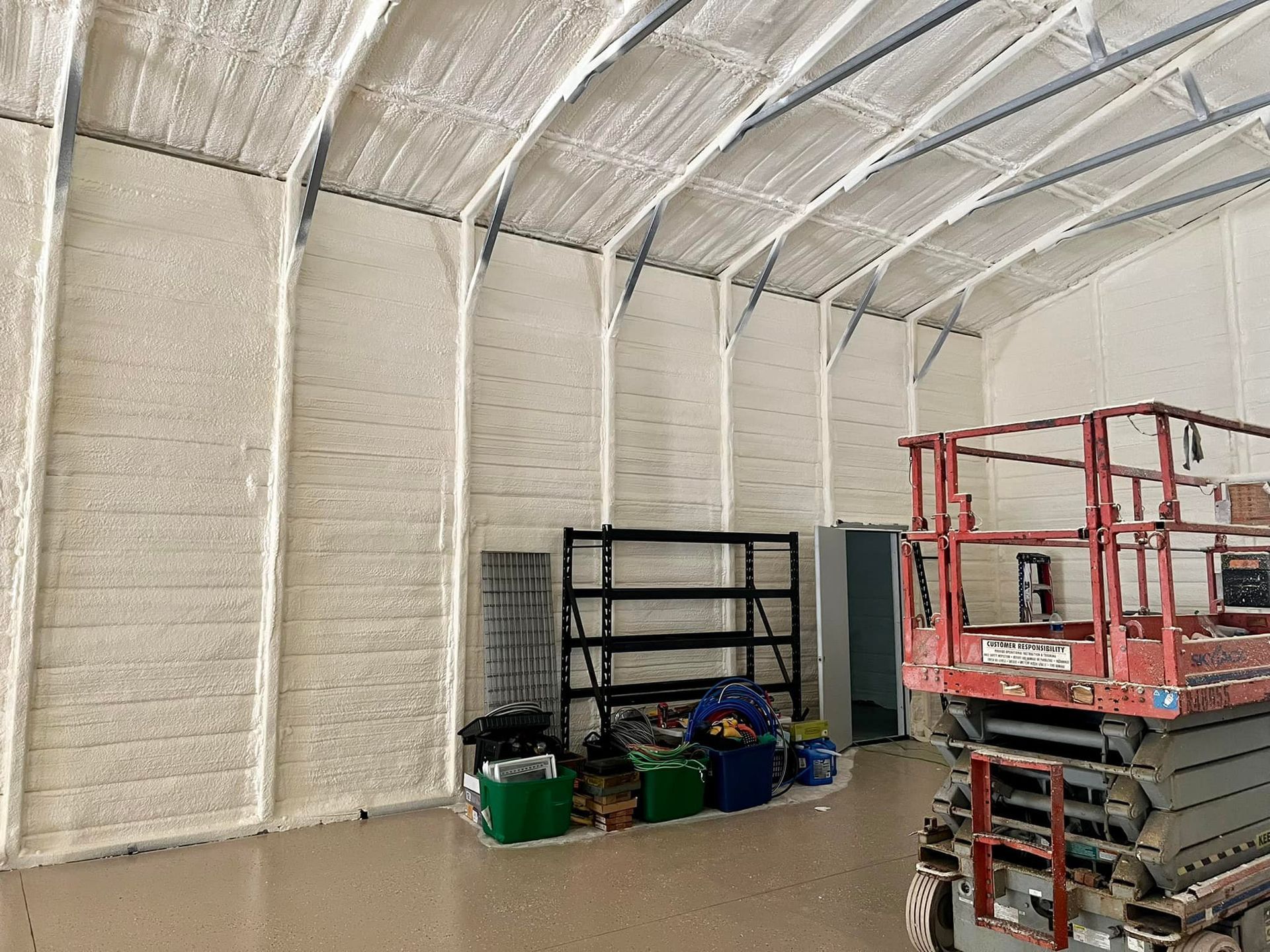 Interior of a large warehouse with spray foam insulation on the walls and ceiling.  A scissor lift and shelving units are visible.