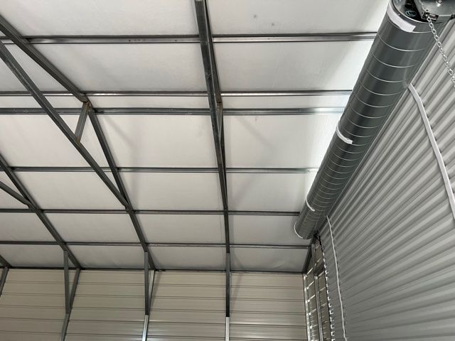 Interior view of a building with a white fabric ceiling supported by a metal frame. A corrugated wall and HVAC duct are visible.