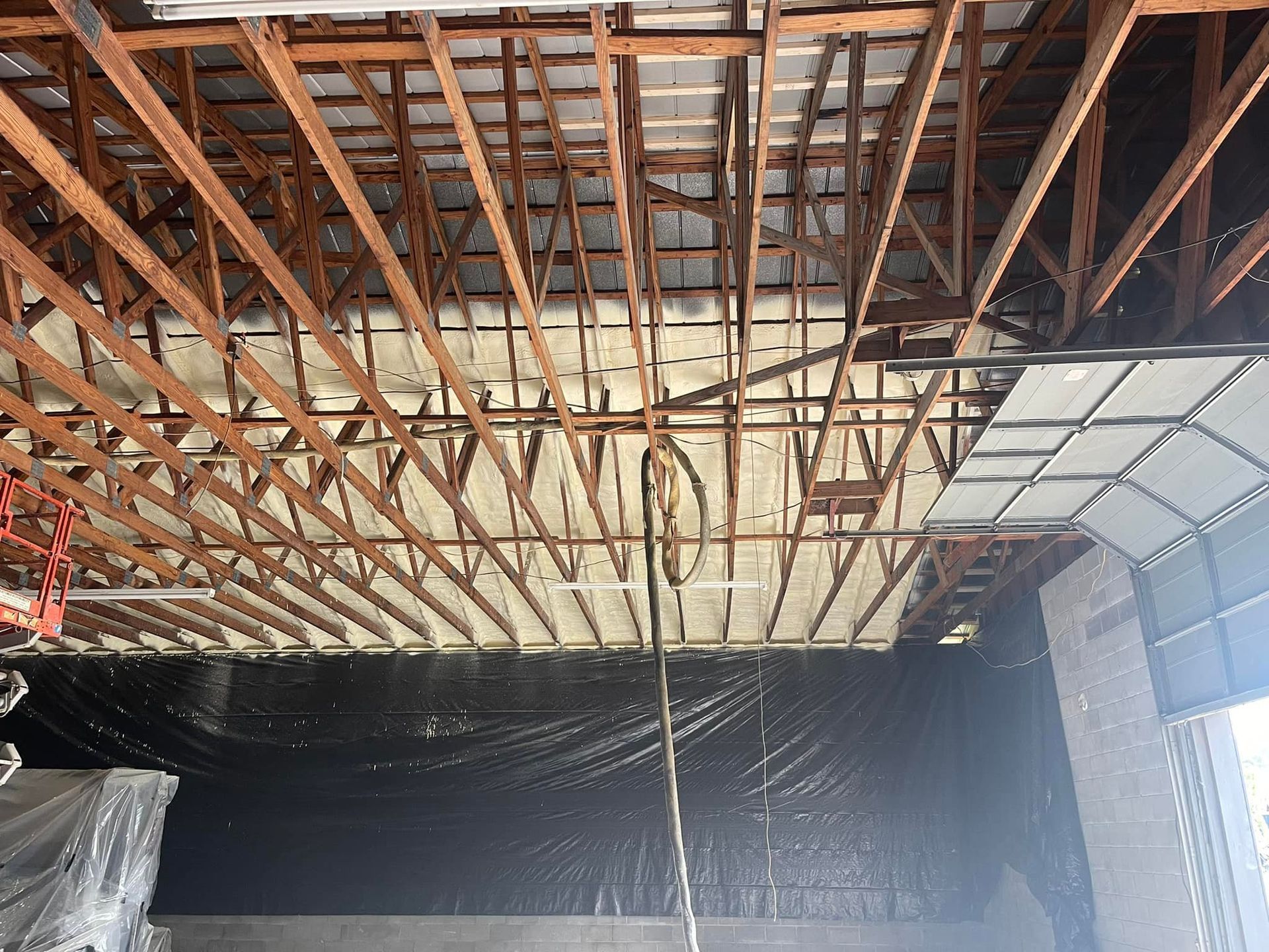 Insulation being sprayed on a warehouse ceiling with wooden beams. Beige foam is applied between the beams.