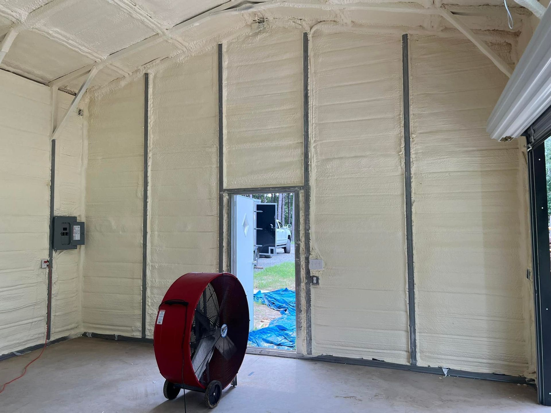 Interior of a structure with spray foam insulation, a large fan, and a partially open door. Beige and grey tones.
