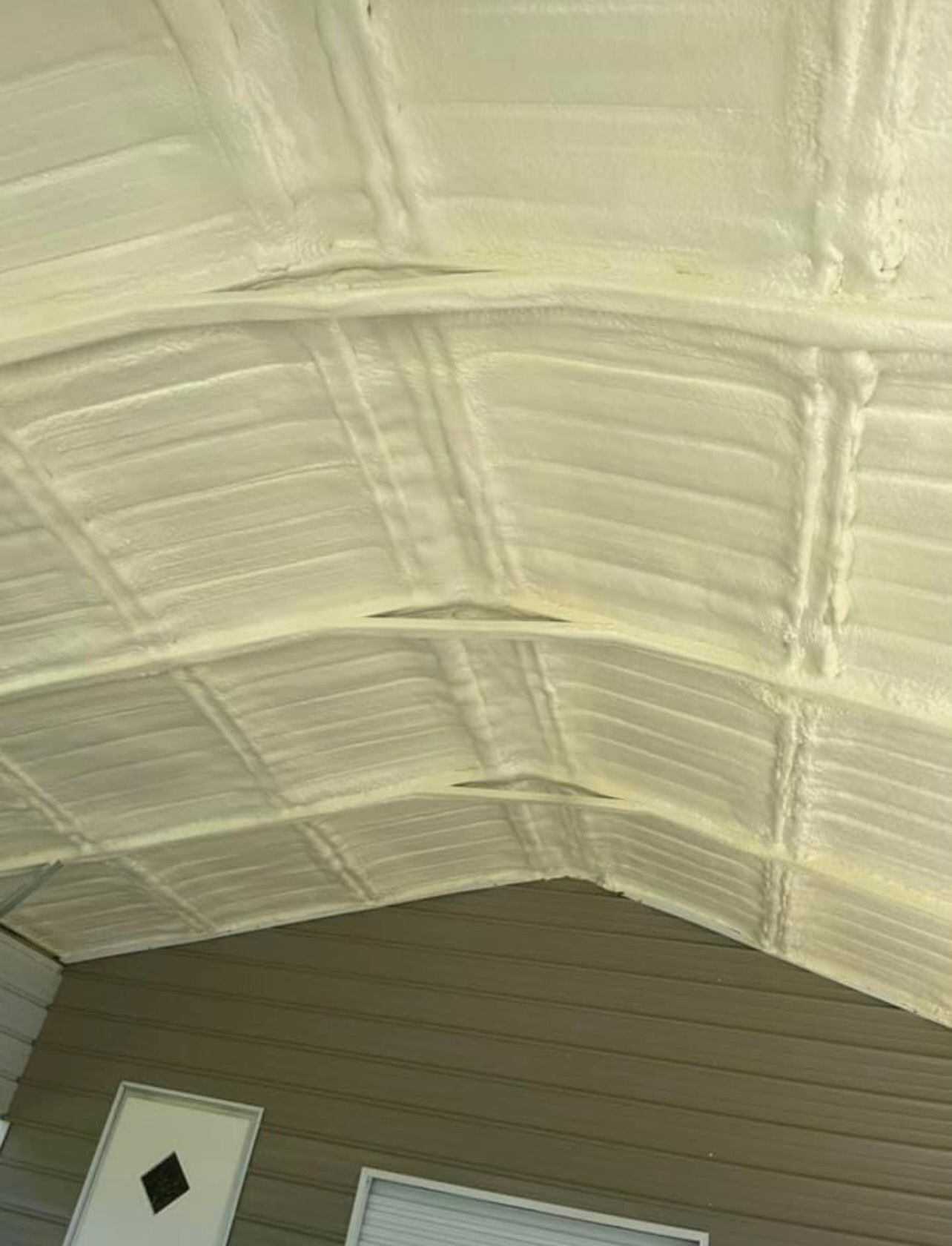 Ceiling insulated with light-colored spray foam, showing beams and a portion of a house exterior.