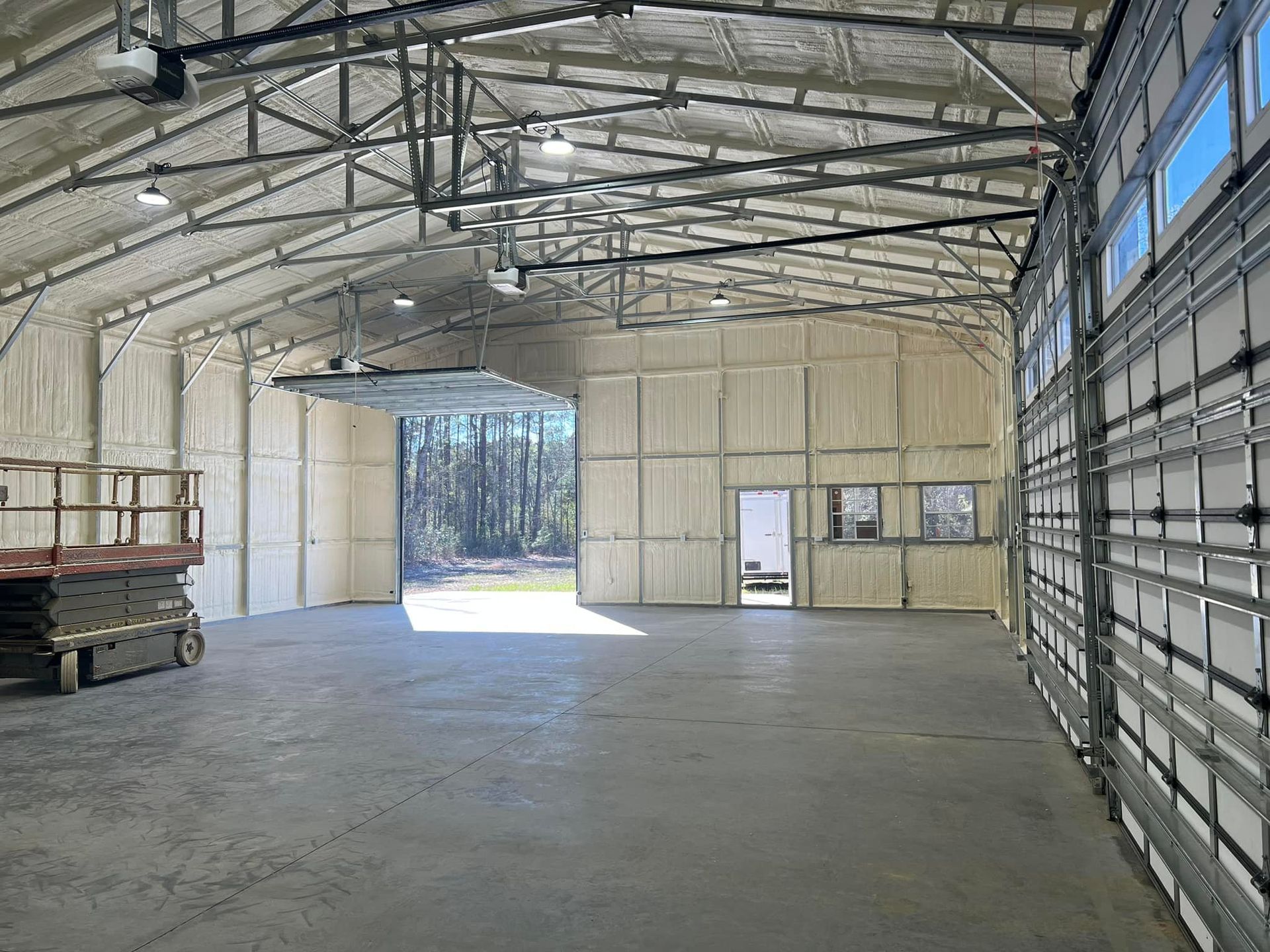 Interior of a large metal building with open doors, concrete floor, and foam insulation. Sunlight streams in.