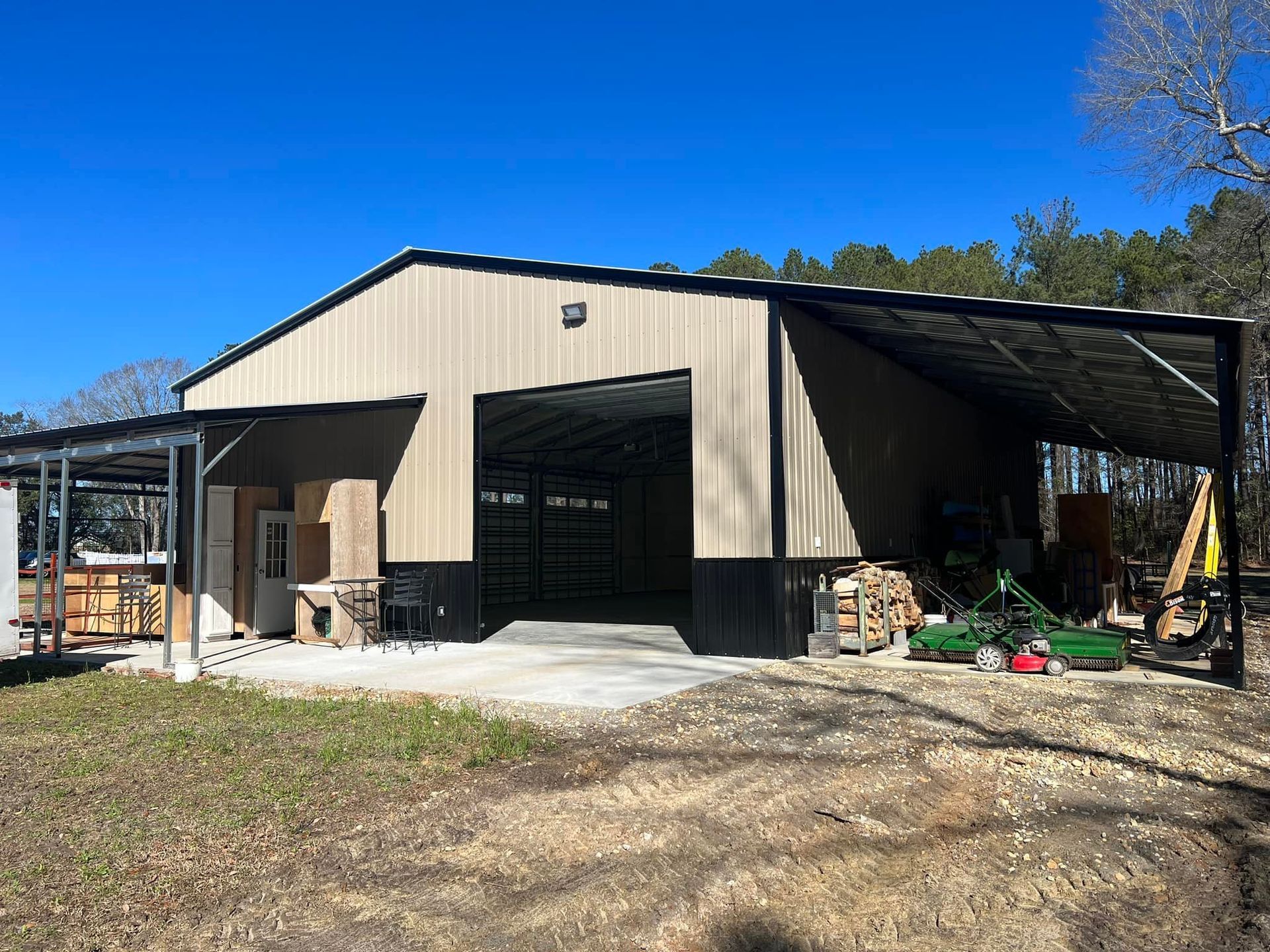 Tan and black metal barn with open bay door and attached canopies; outdoors on a sunny day.