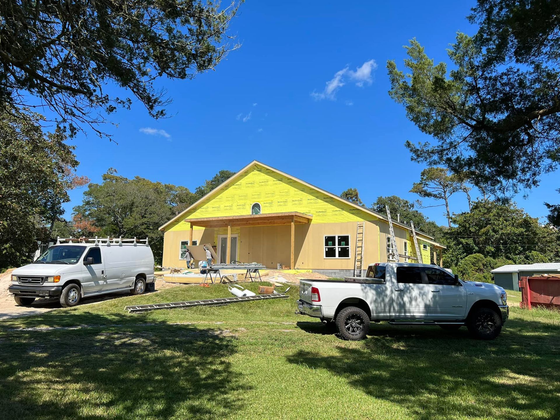 Construction site: partially built house with two work trucks and surrounding trees. Blue sky.