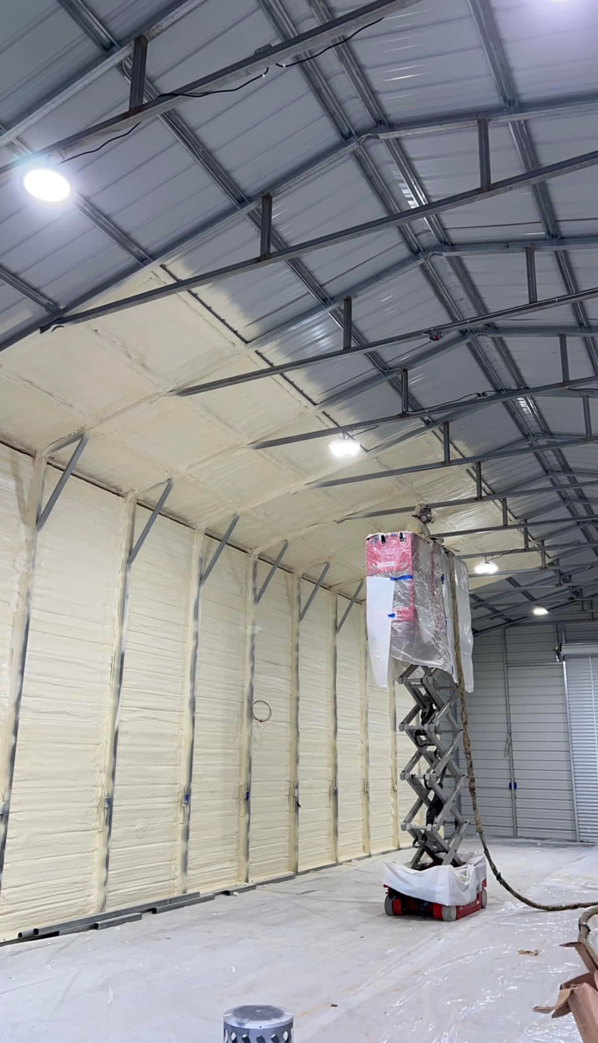 Interior of a building with white foam insulation on the walls and ceiling, a scissor lift, and bright overhead lights.
