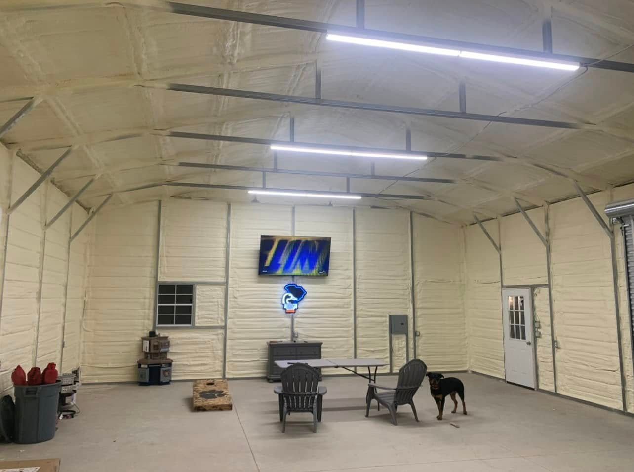 Inside a garage: spray foam insulation, TV, seating, dog, and a cornhole game.