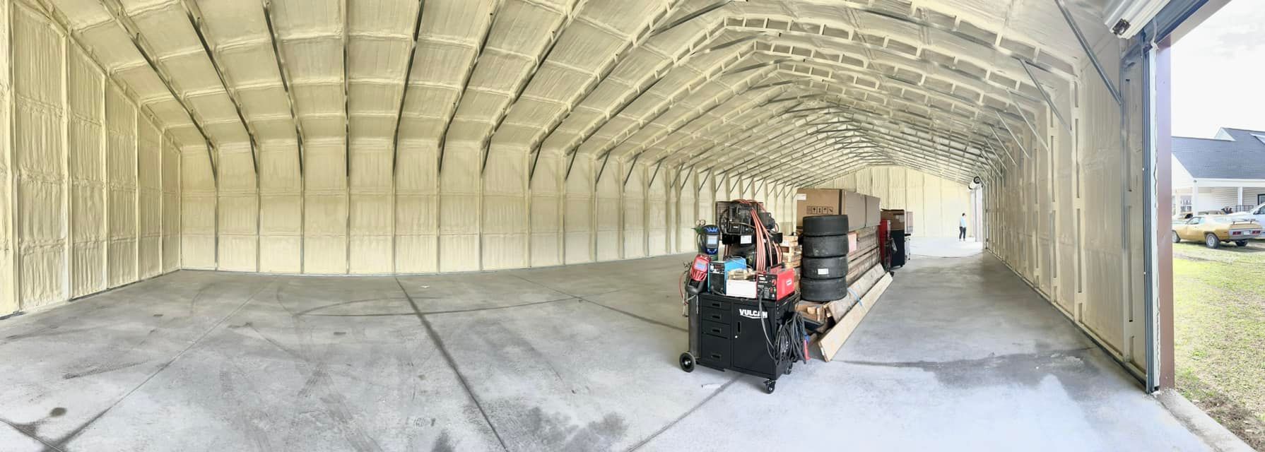 Interior of a wide, empty building with spray foam insulation. A garage door is open to a grassy yard.