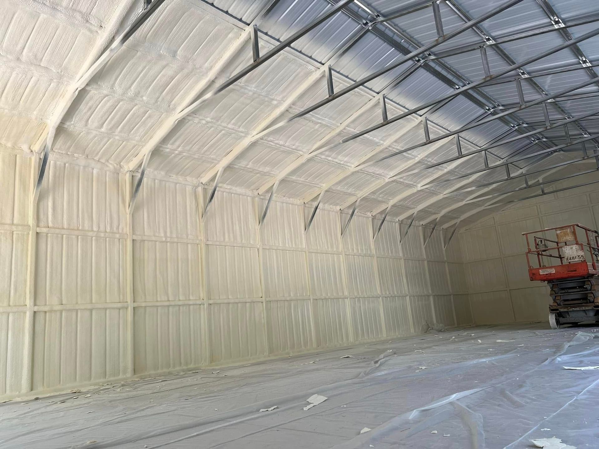 Interior of a metal building insulated with light tan spray foam. A lift is in the corner.