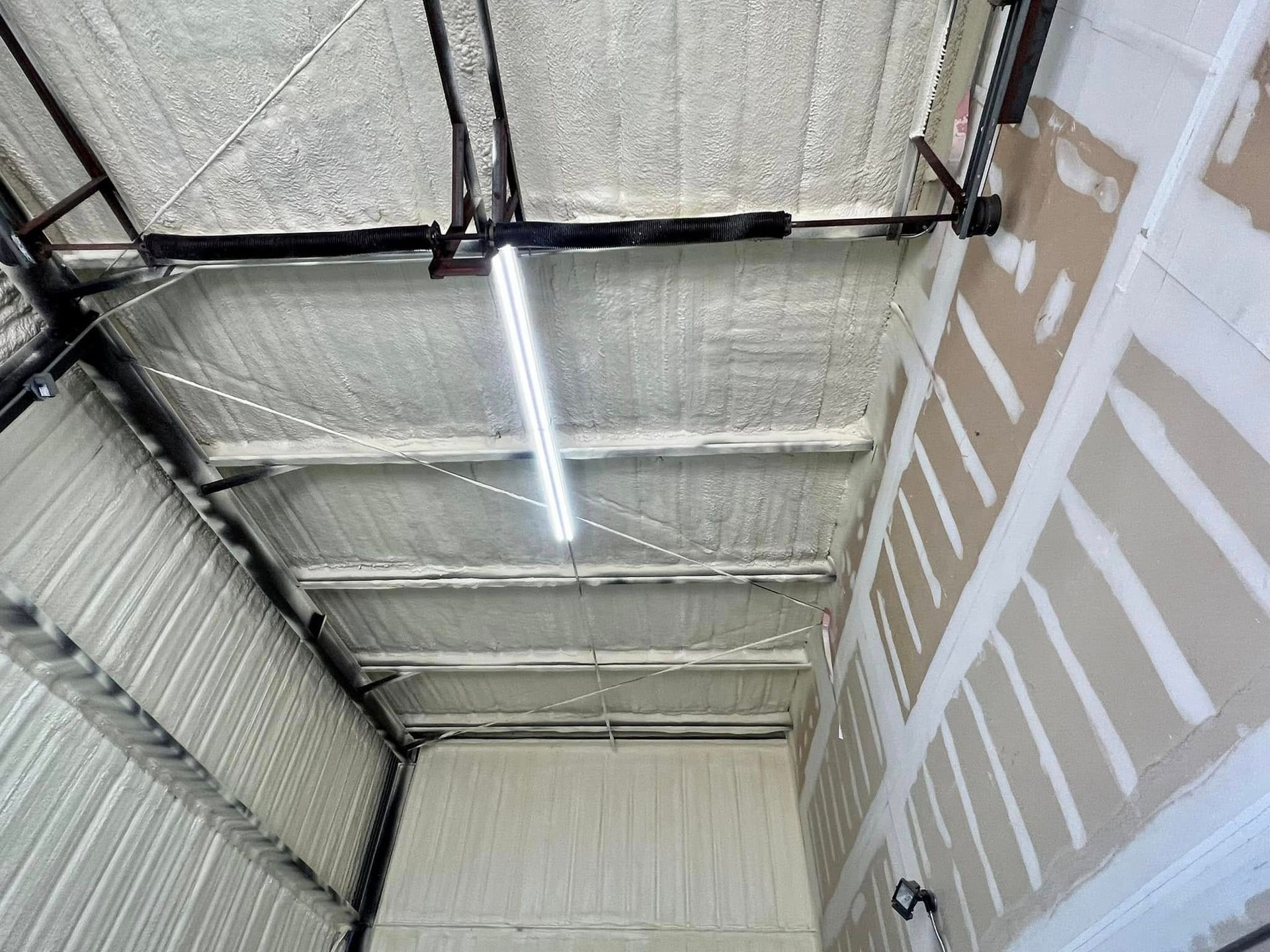 Interior view of a garage with spray foam insulation on the roof and walls. A fluorescent light hangs down.