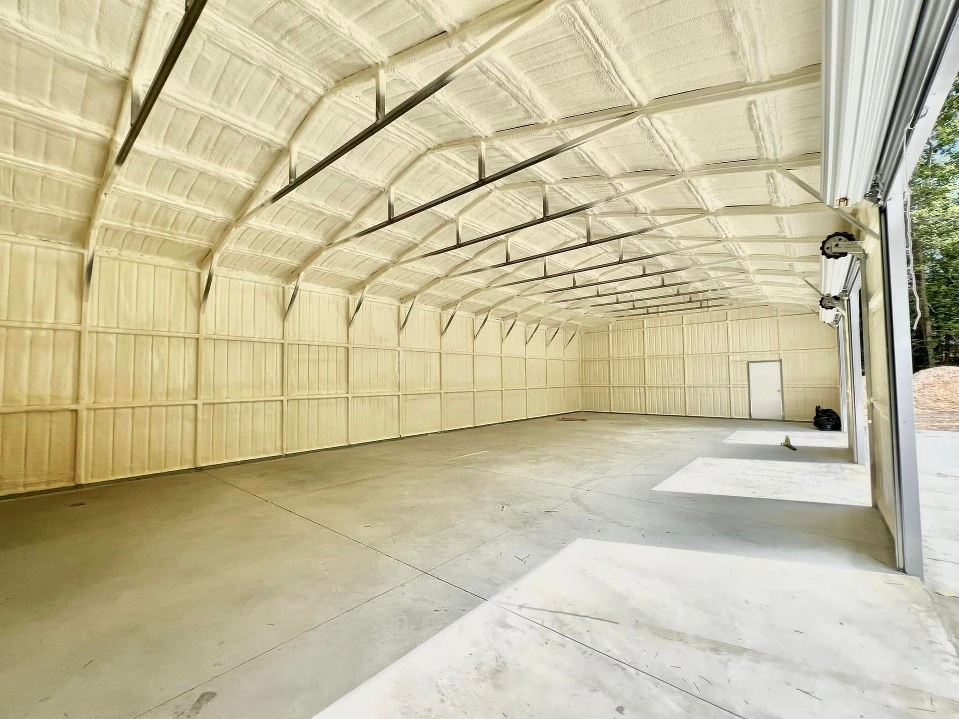 Interior of a large, empty warehouse with spray foam insulation on walls and ceiling. Concrete floor, garage door visible.
