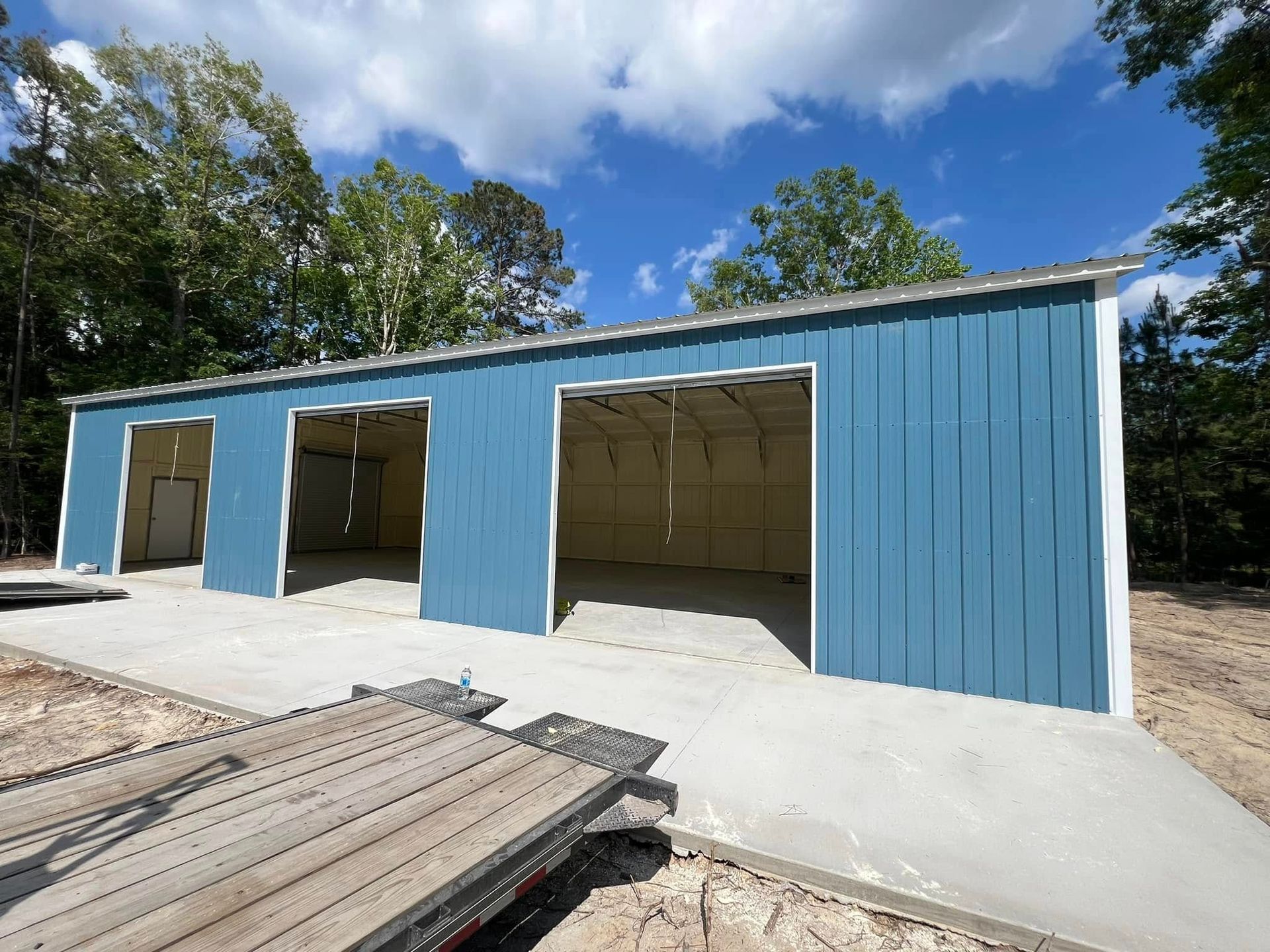 Blue metal garage with three bays and concrete foundation on a sunny day.