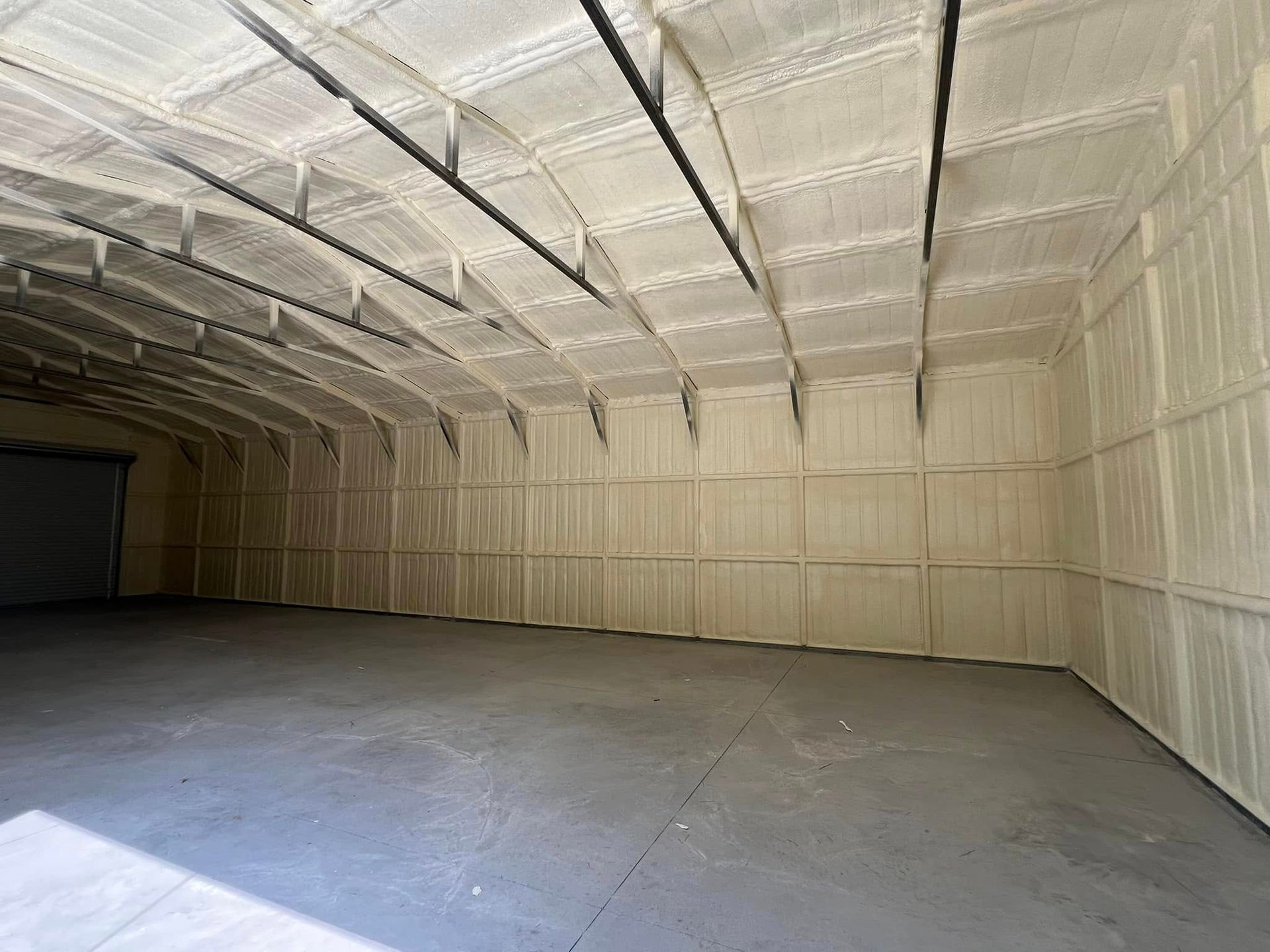 Interior of a large, empty warehouse; walls and ceiling covered in beige spray foam insulation, concrete floor.