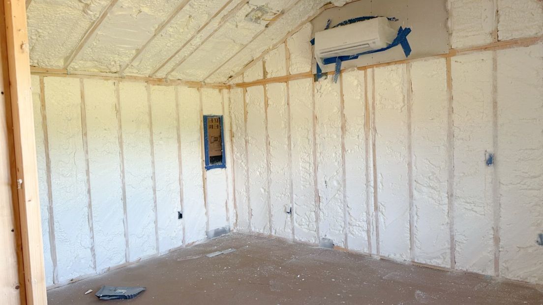 Interior of a room with spray foam insulation between wooden studs, and an air conditioning unit.