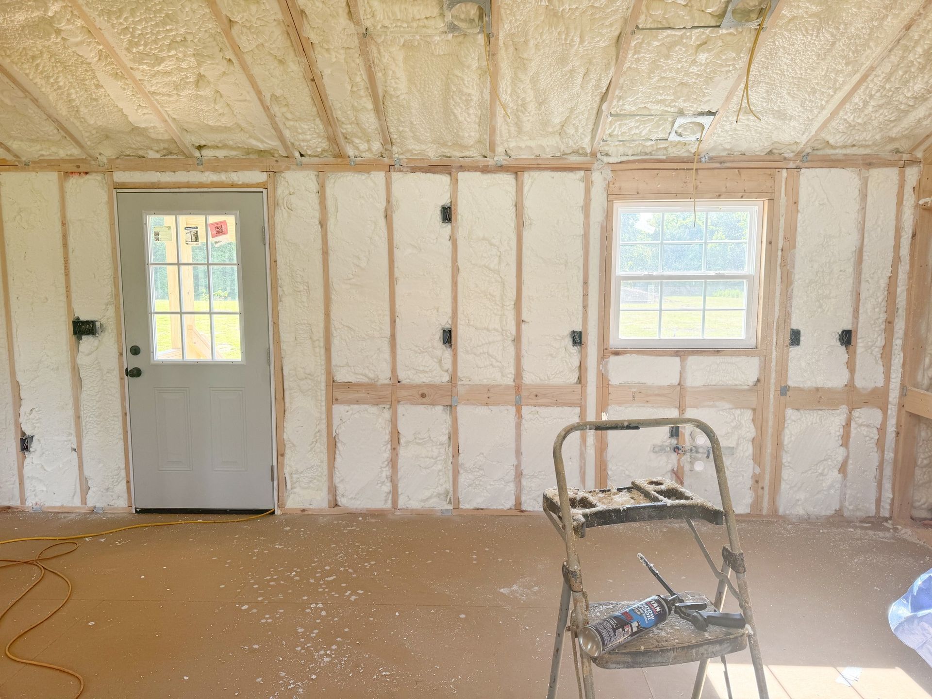 Interior of a room with spray foam insulation on walls and ceiling; a door, window, and step ladder present.