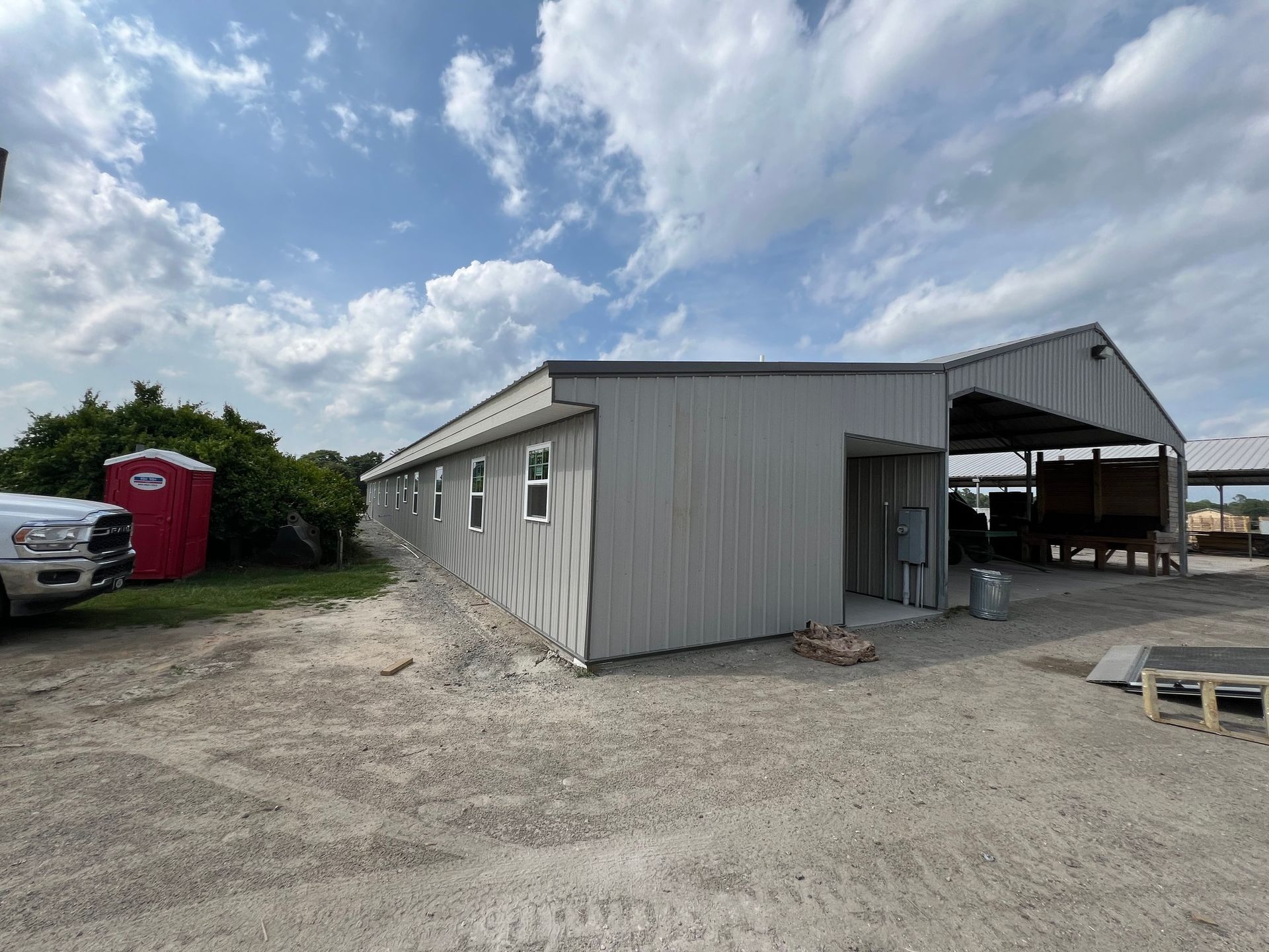 Gray metal building with open-air shelter attached; construction site with porta-potty and cloudy sky.