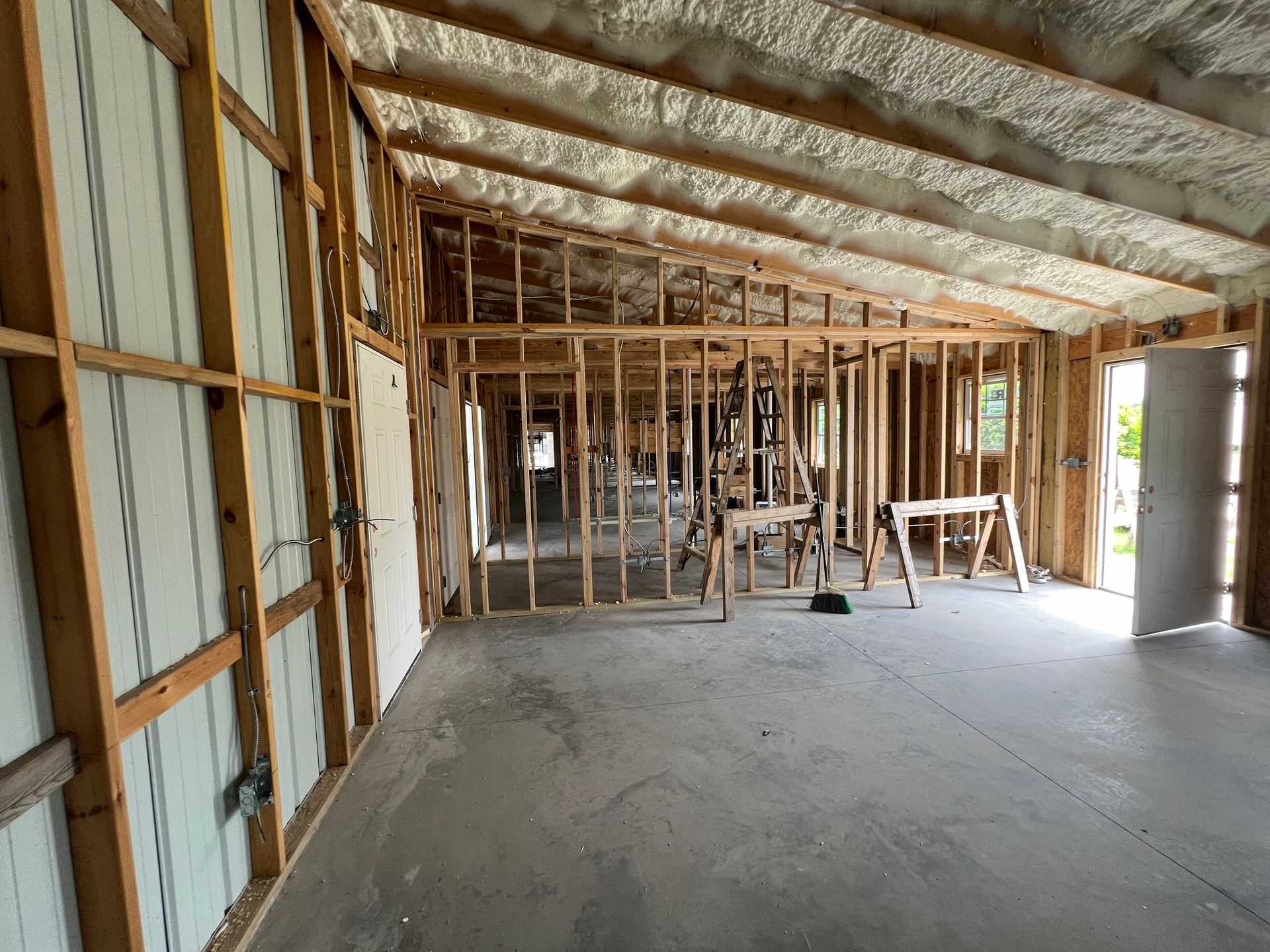 Interior view of a building under construction, showing wooden frames, concrete floor, and spray foam insulation.