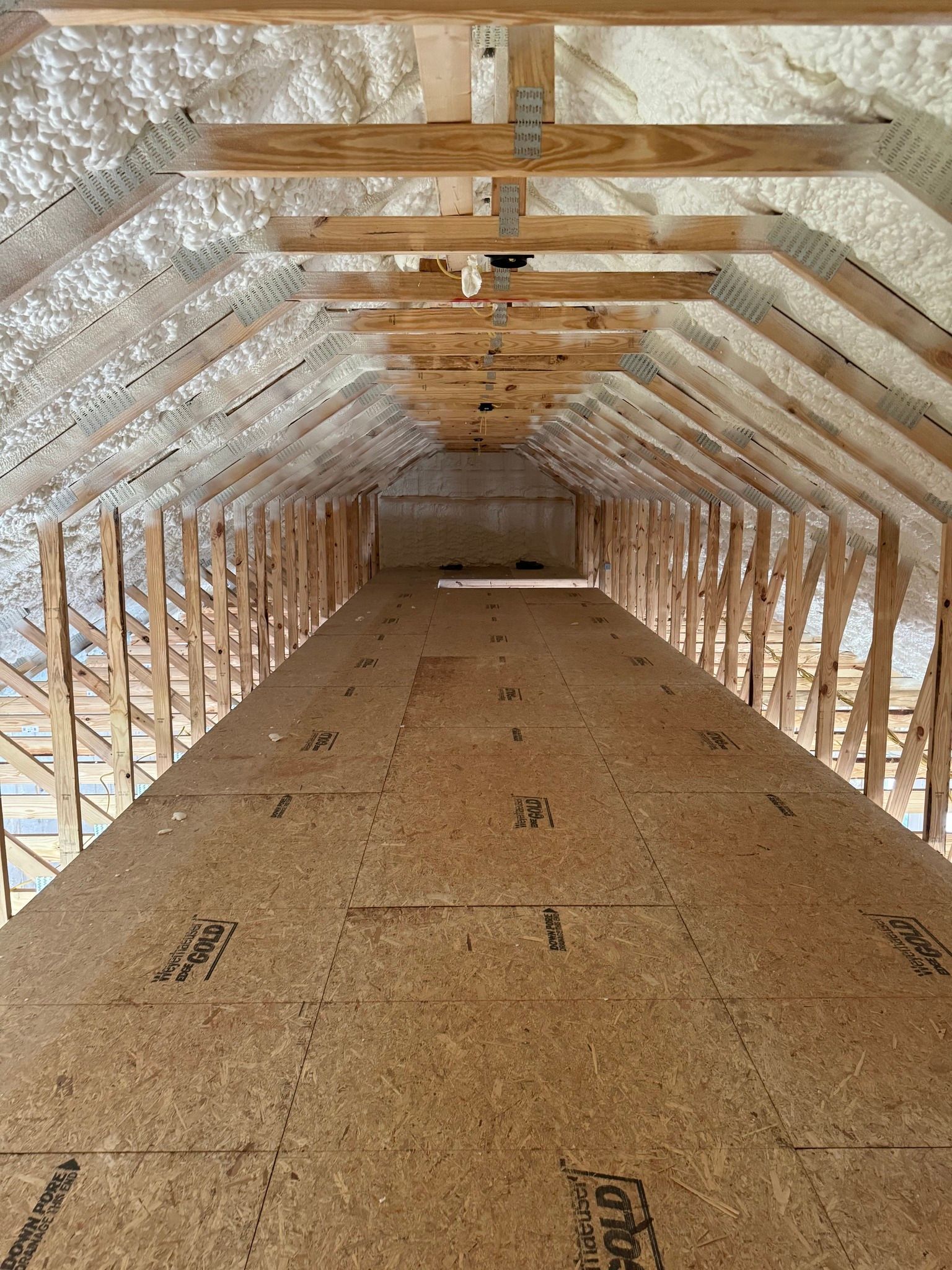 Attic interior with plywood floor and spray foam insulation on the rafters. Wooden beams are visible.