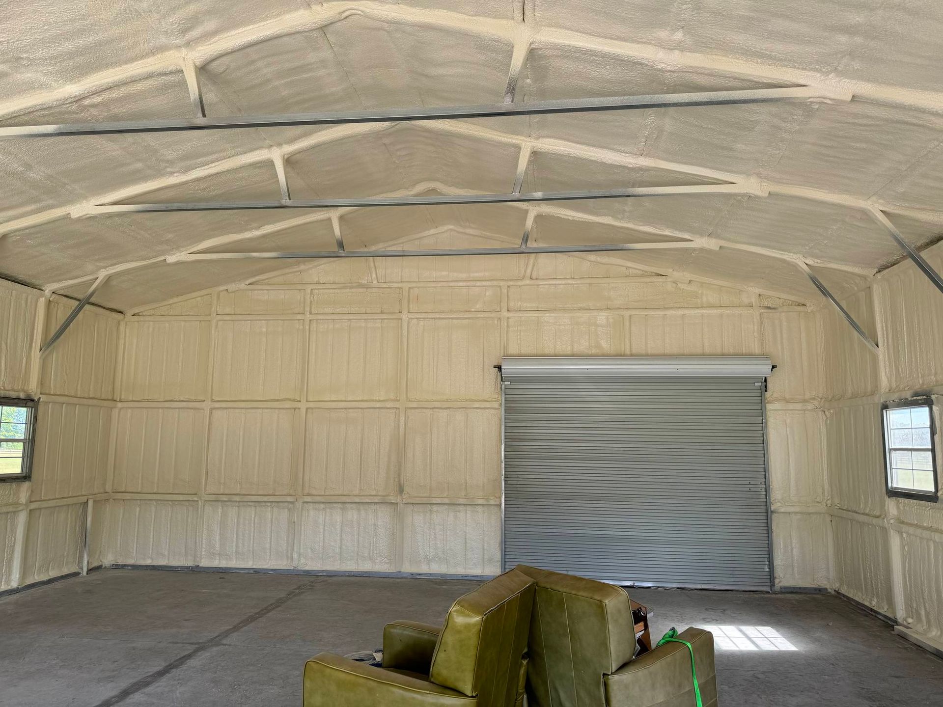 Interior of a building with spray foam insulation on walls and ceiling, gray garage door, windows.