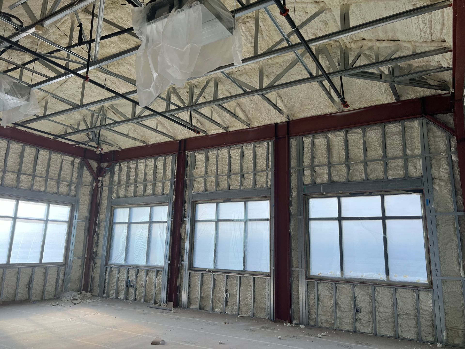 Interior of a building under construction, showing insulated walls, steel beams, and windows. The ceiling is also insulated.