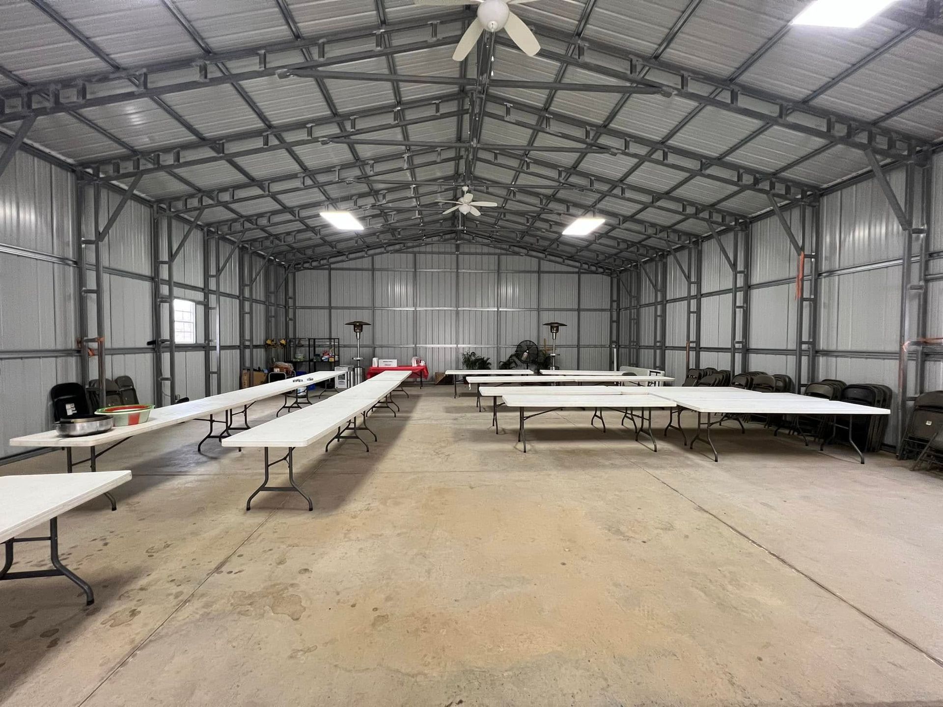 Interior of a large metal building with white folding tables set up. Concrete floor, ceiling fans.