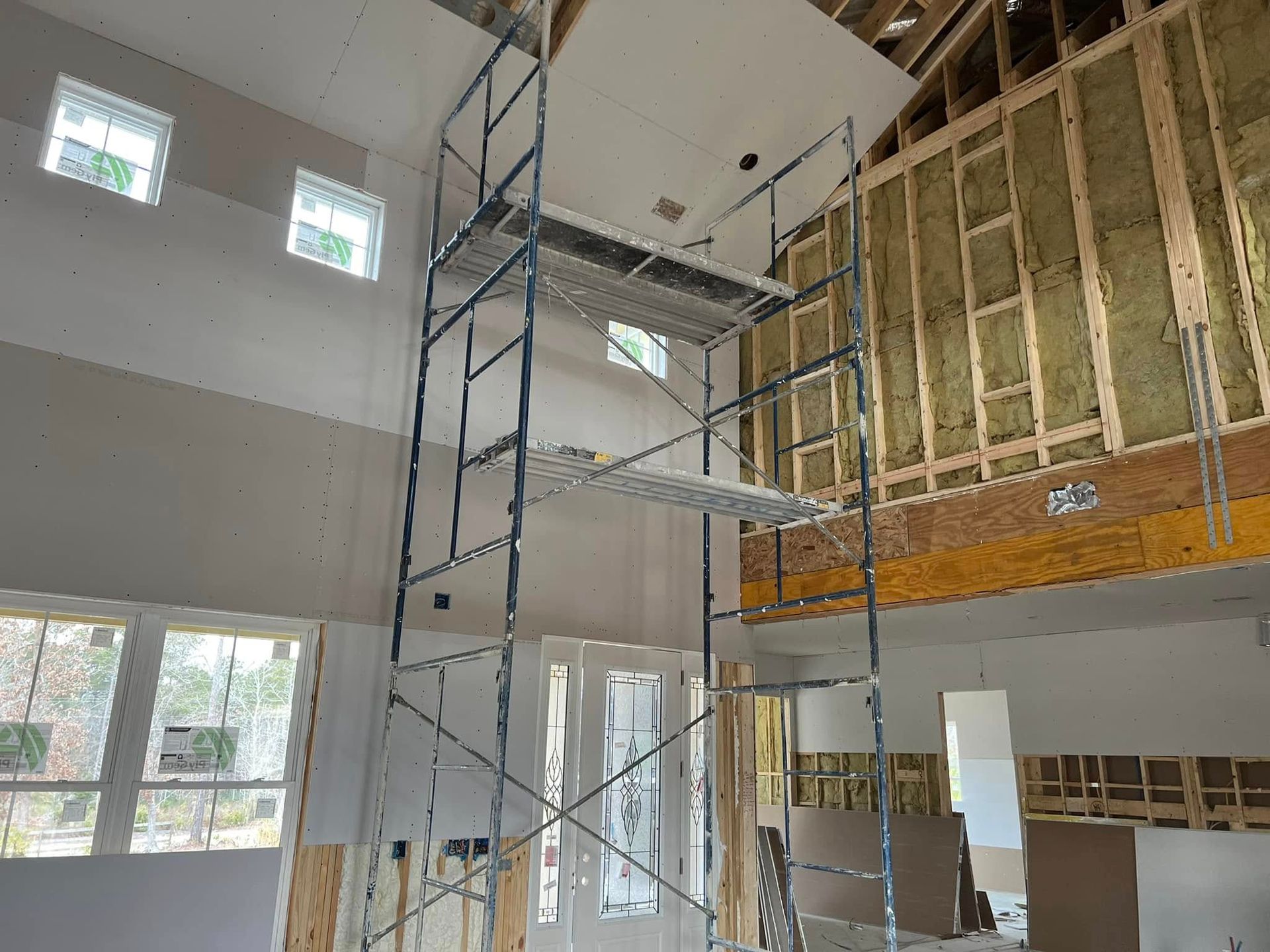 Scaffolding inside a building under construction, reaching a high ceiling. Drywall is being installed.