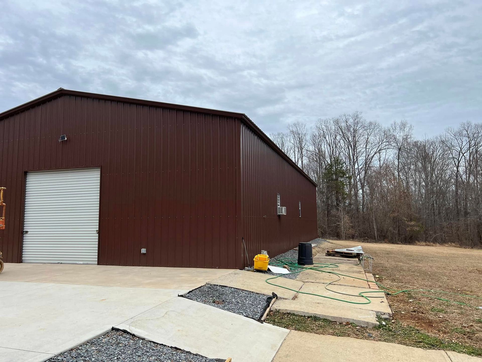 Brown metal building with a white garage door, concrete pad, and trees in the background under a cloudy sky.