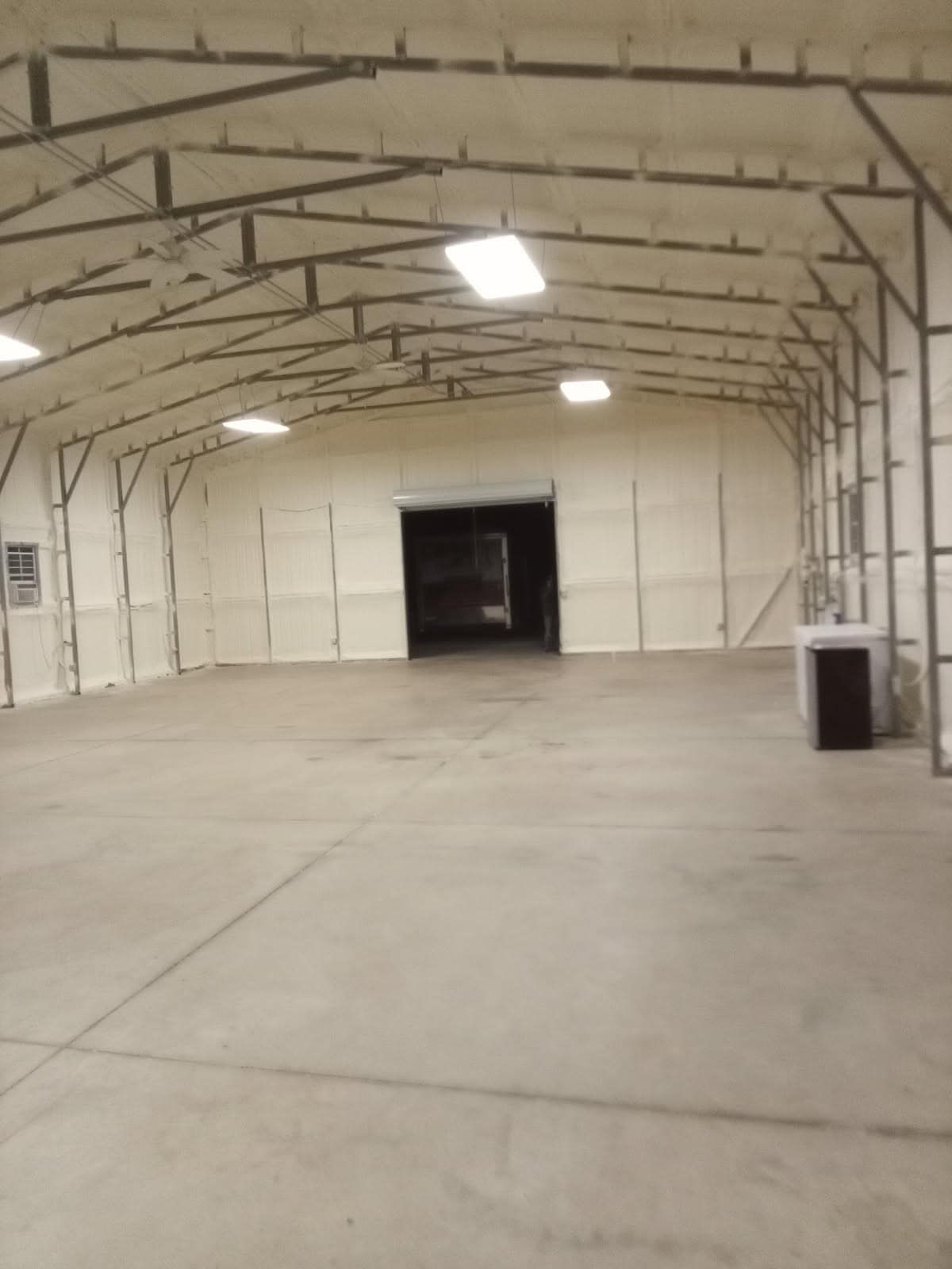 Empty warehouse interior, gray concrete floor, white insulated walls, steel framework, overhead lights, open doorway.