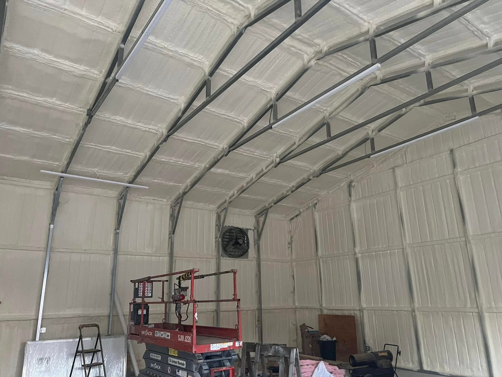 Interior of a building with curved metal framing and spray foam insulation on the walls and ceiling. A lift and other equipment are visible.