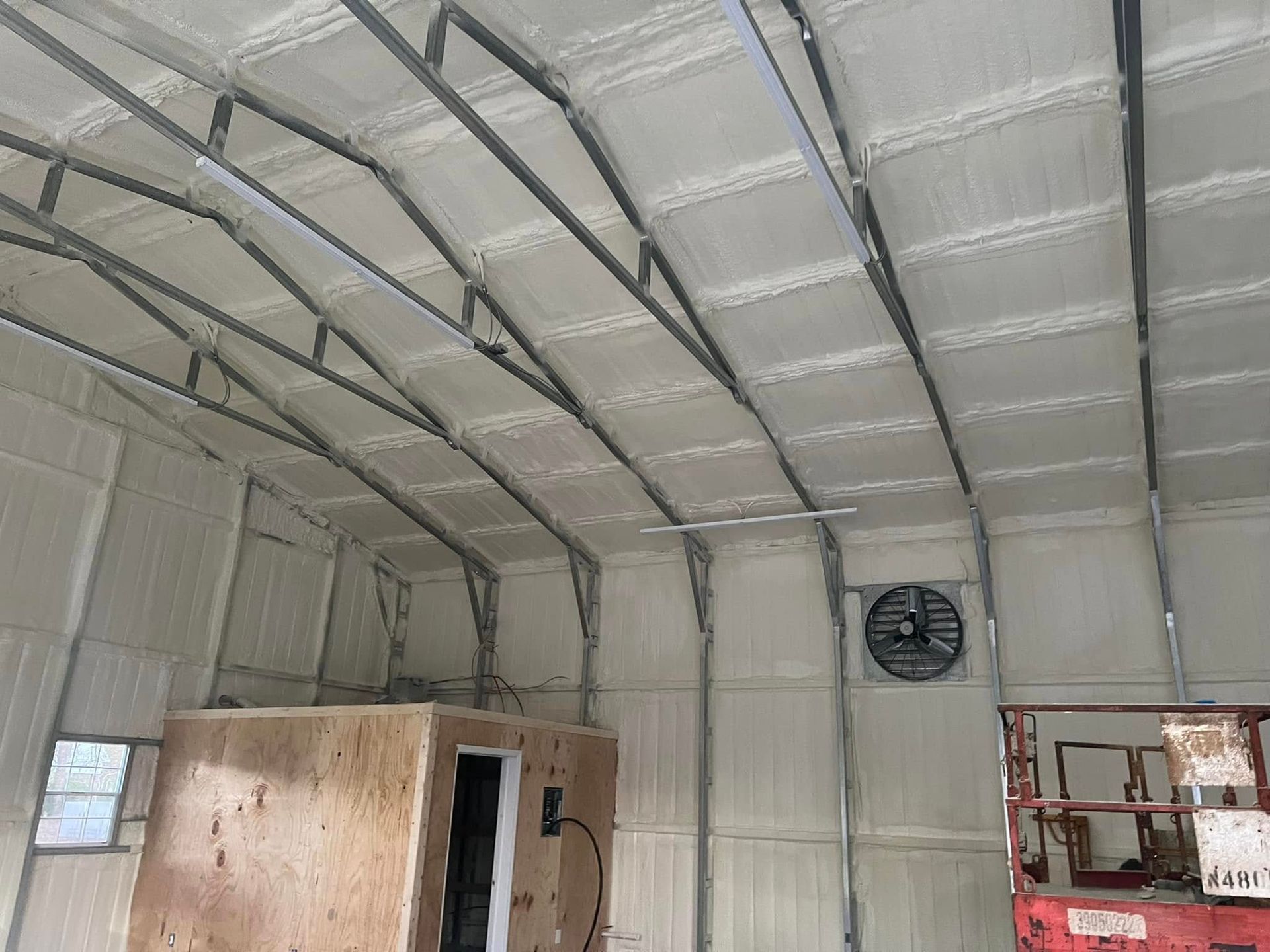 Interior of a metal building with spray foam insulation, including ceiling and walls. Features a fan and a small wooden structure.