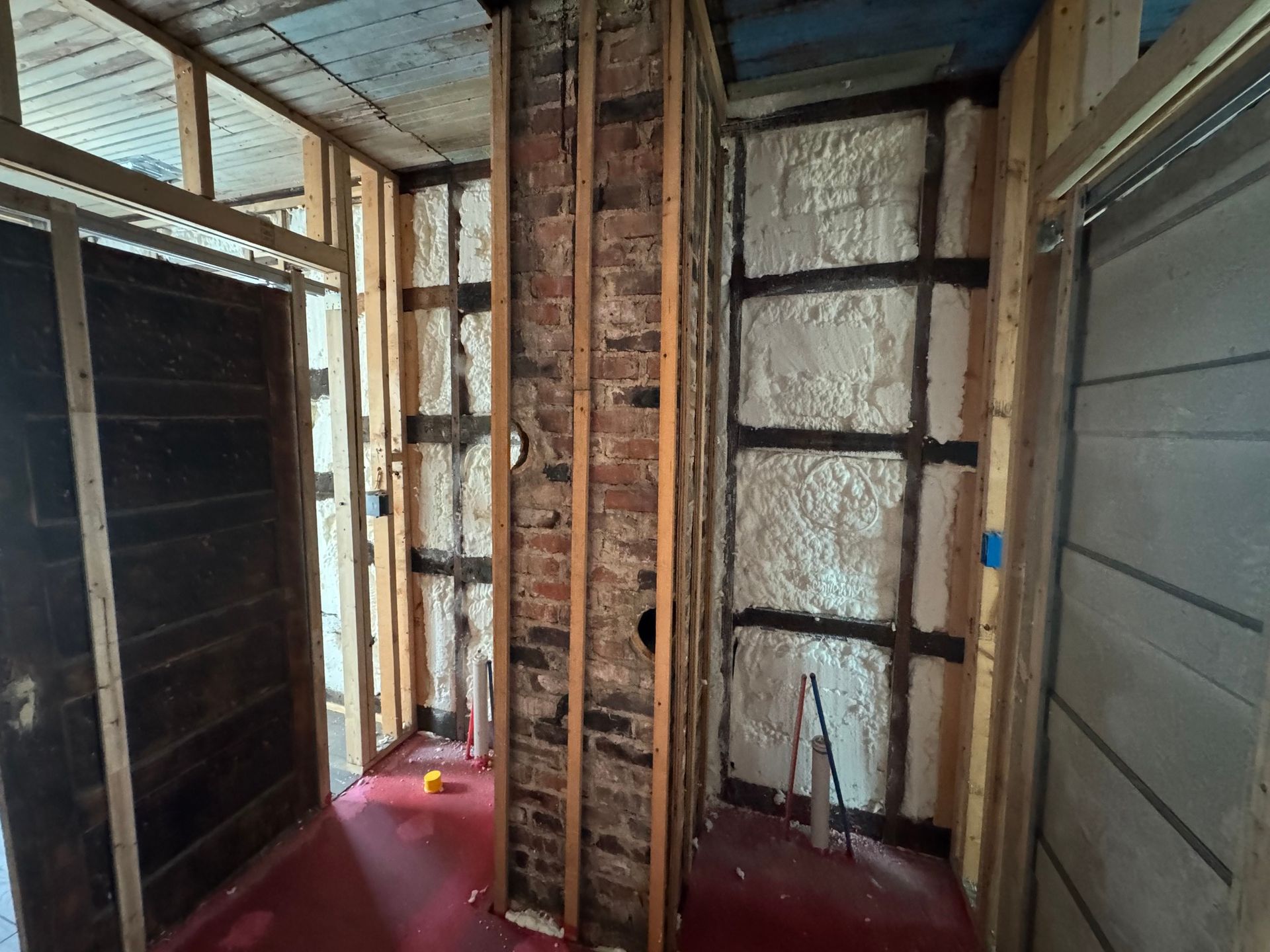 Interior view of a room under construction with exposed brick chimney and spray foam insulation between wooden studs.