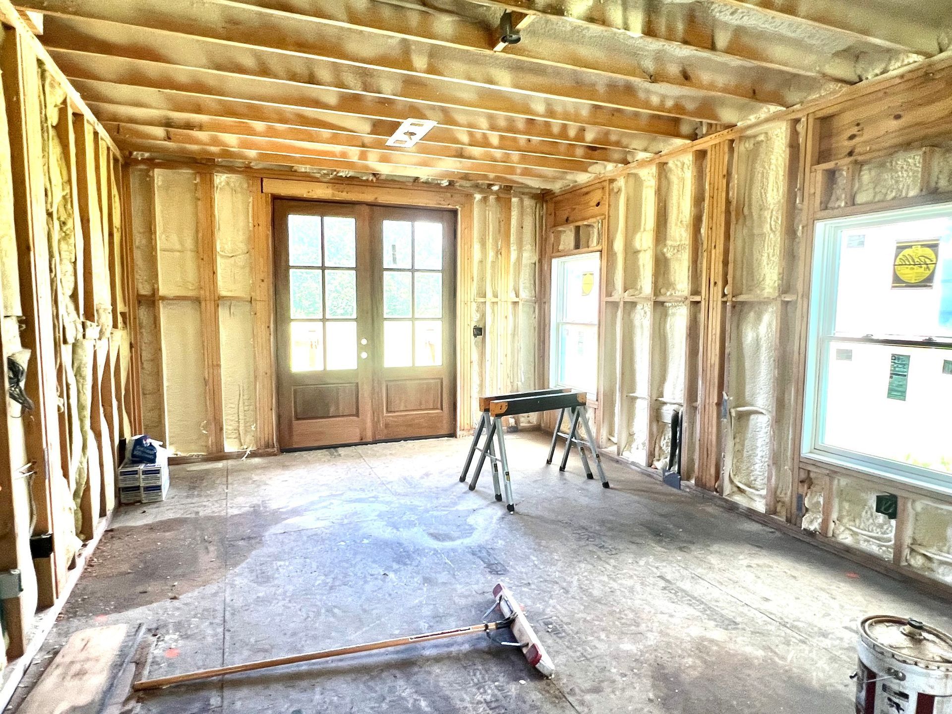 Interior room under construction, with spray foam insulation visible on walls, door, window, and sawhorse.