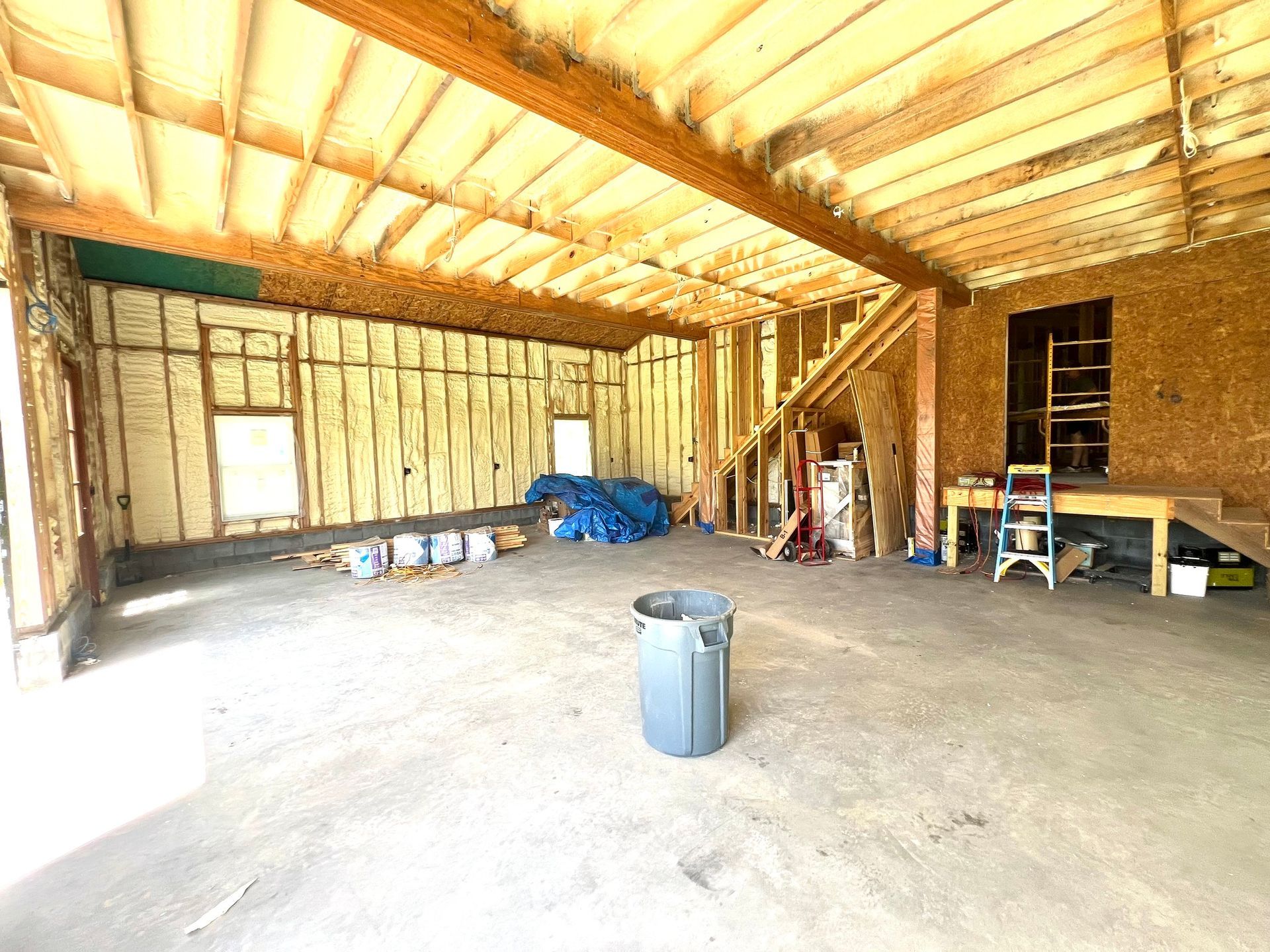 Interior of a garage under construction with exposed wooden beams, insulation, and concrete floor; a trash can sits in the center.