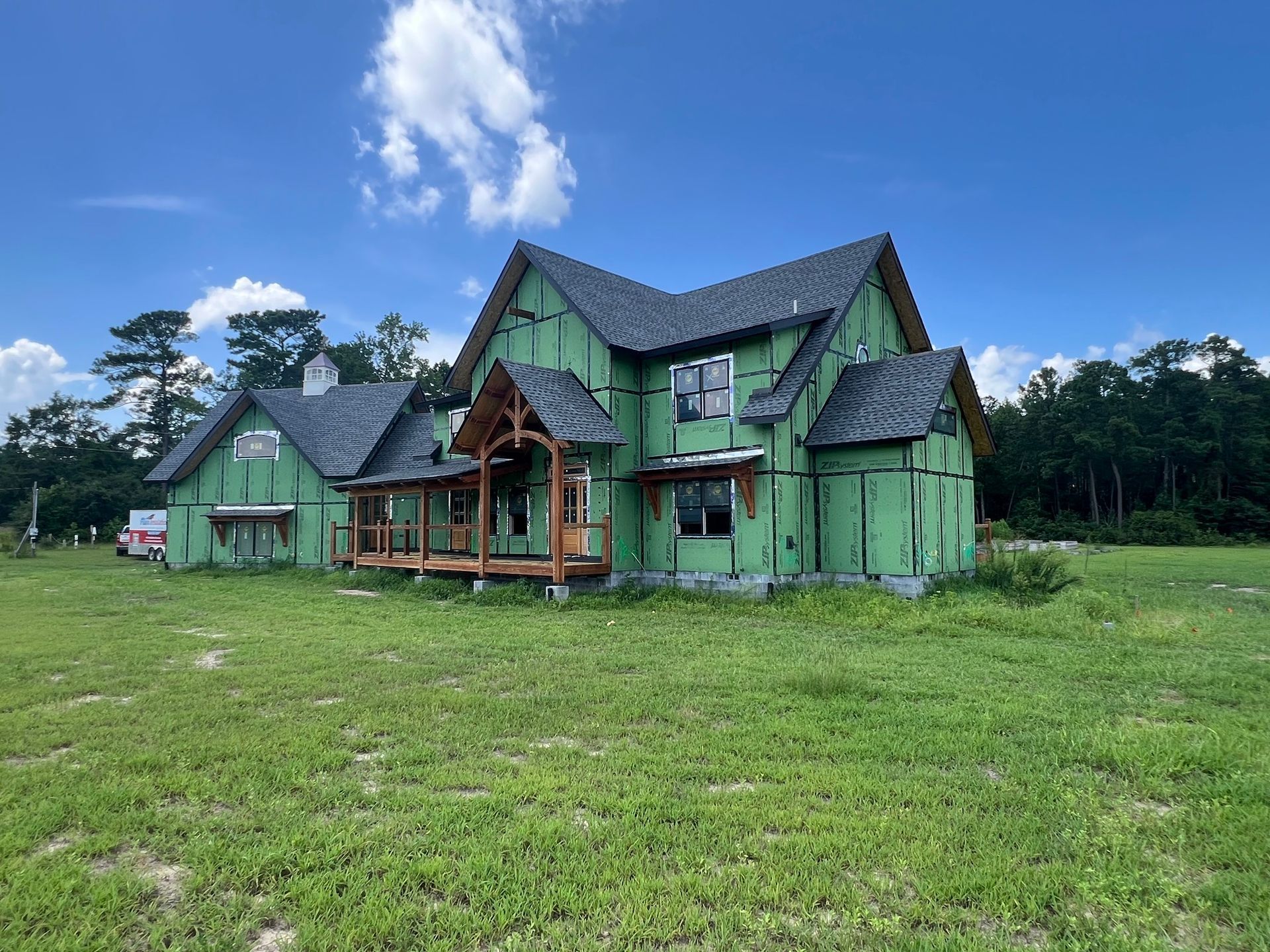 New house under construction with green wrap and dark roof, in a grassy field on a sunny day.
