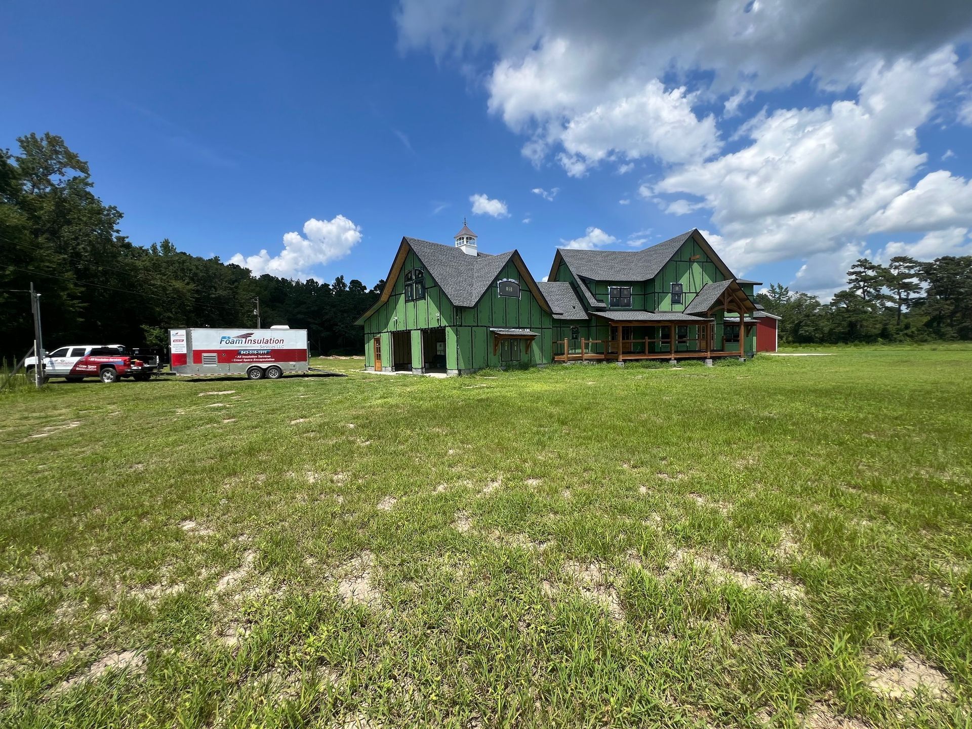 House under construction with green siding, green roof, and a red trailer on a grassy field under a blue sky.