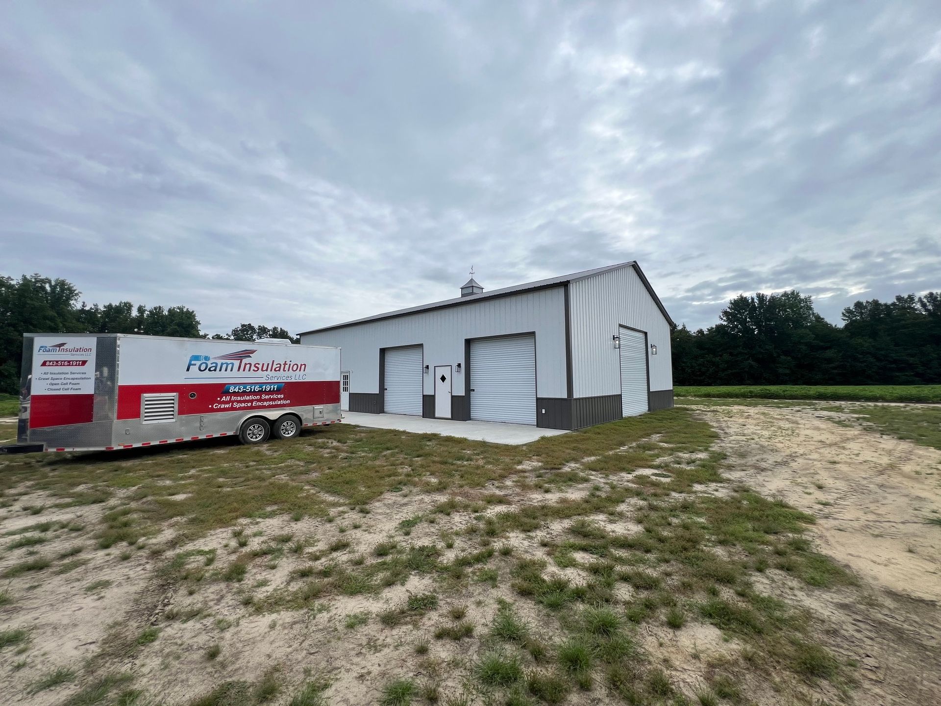 White and gray metal building with two garage doors; red and white trailer on the left; cloudy sky.